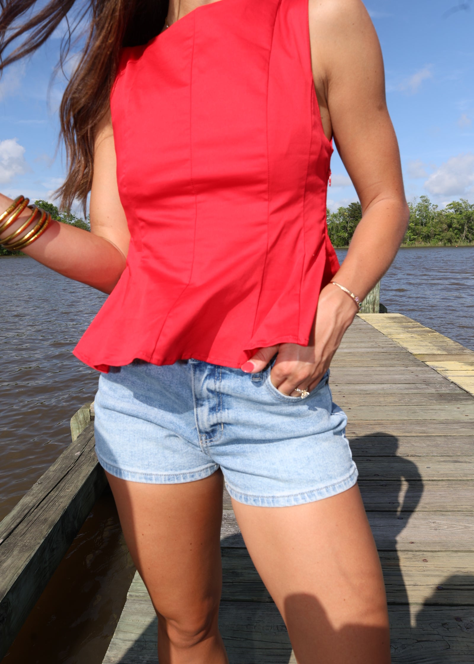 Person wearing a red top and denim shorts standing on a wooden deck with a blue sky background.