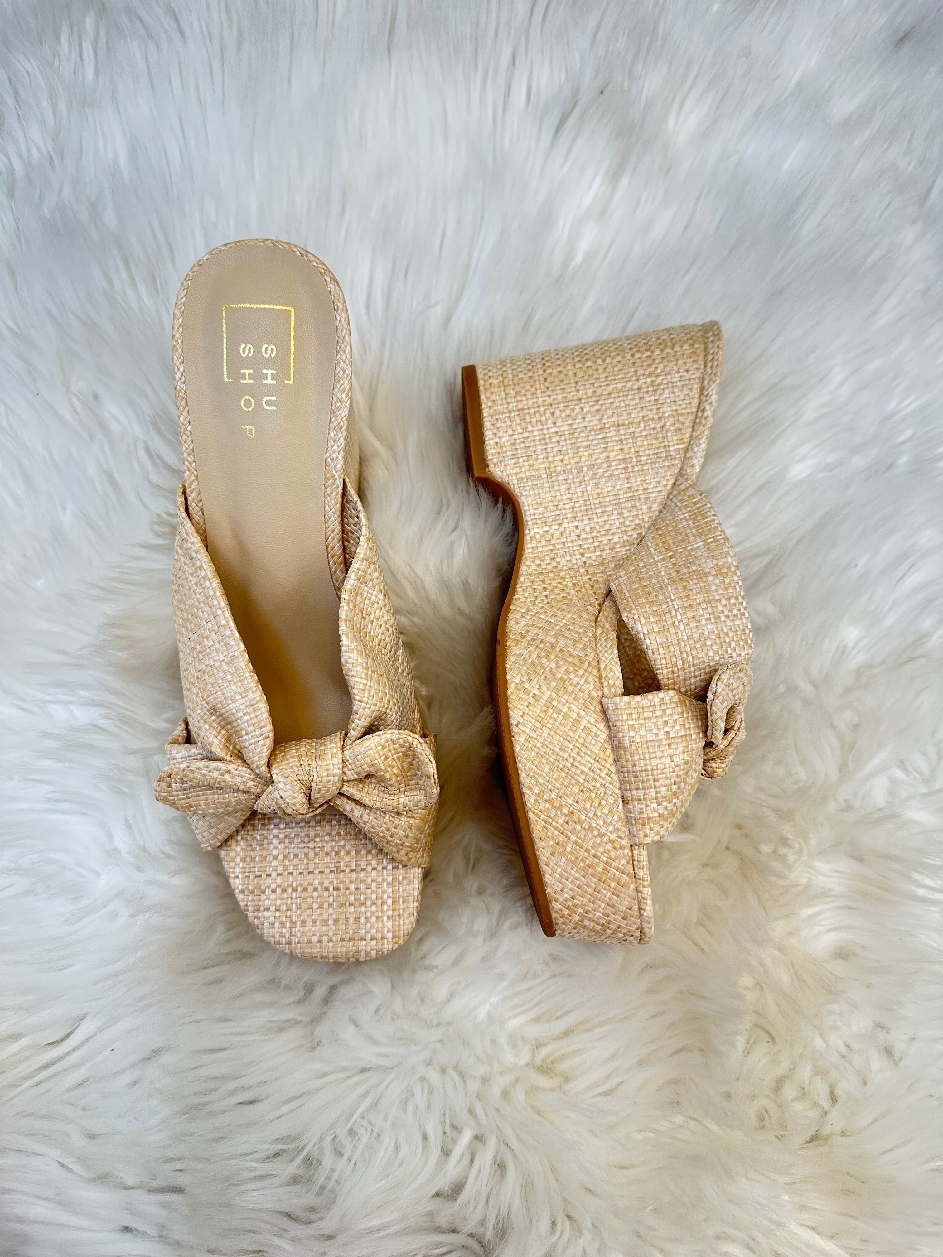 Pair of beige woven wedge sandals with a bow on a white fur background