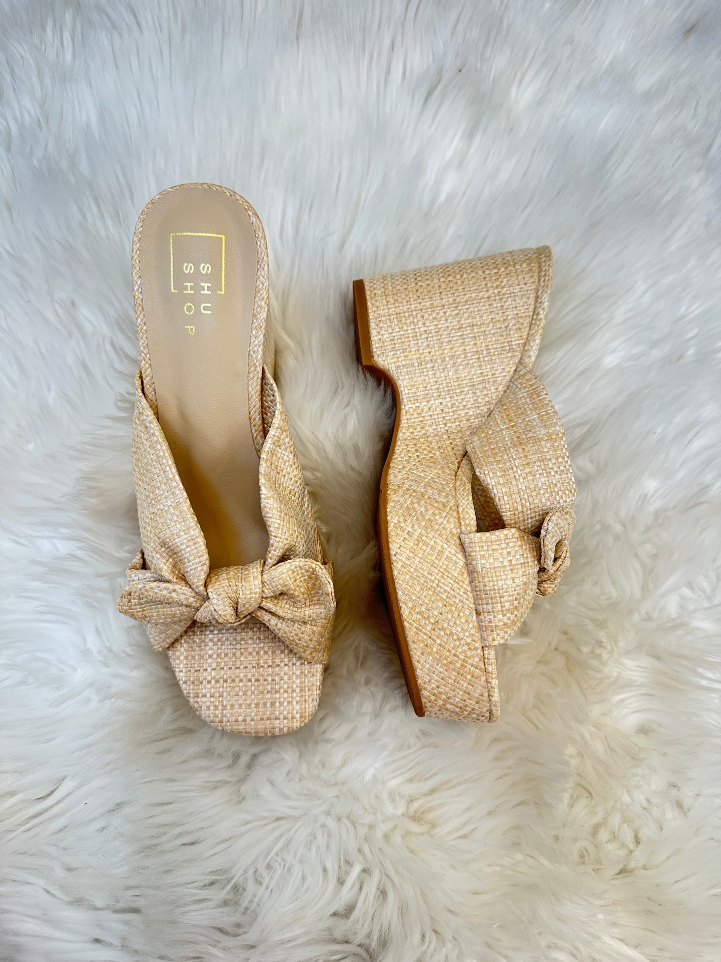 Pair of beige woven wedge sandals with a bow on a white fur background
