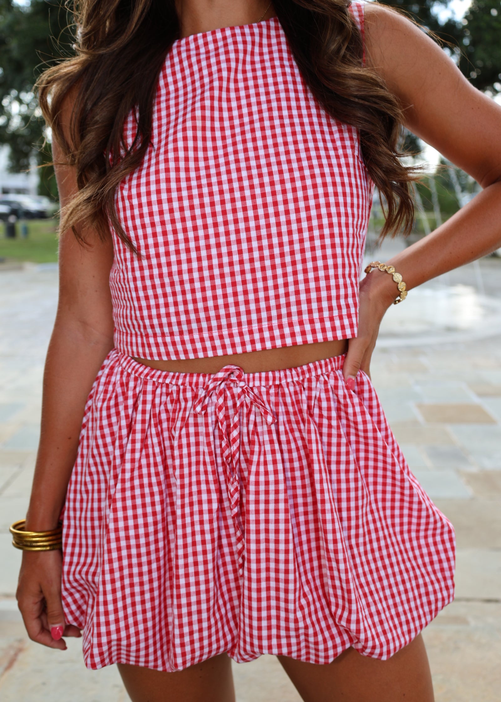 A woman wearing a red and white gingham check two-piece set consisting of a sleeveless crop top and matching ruffled shorts, shown in a close-up lifestyle shot.