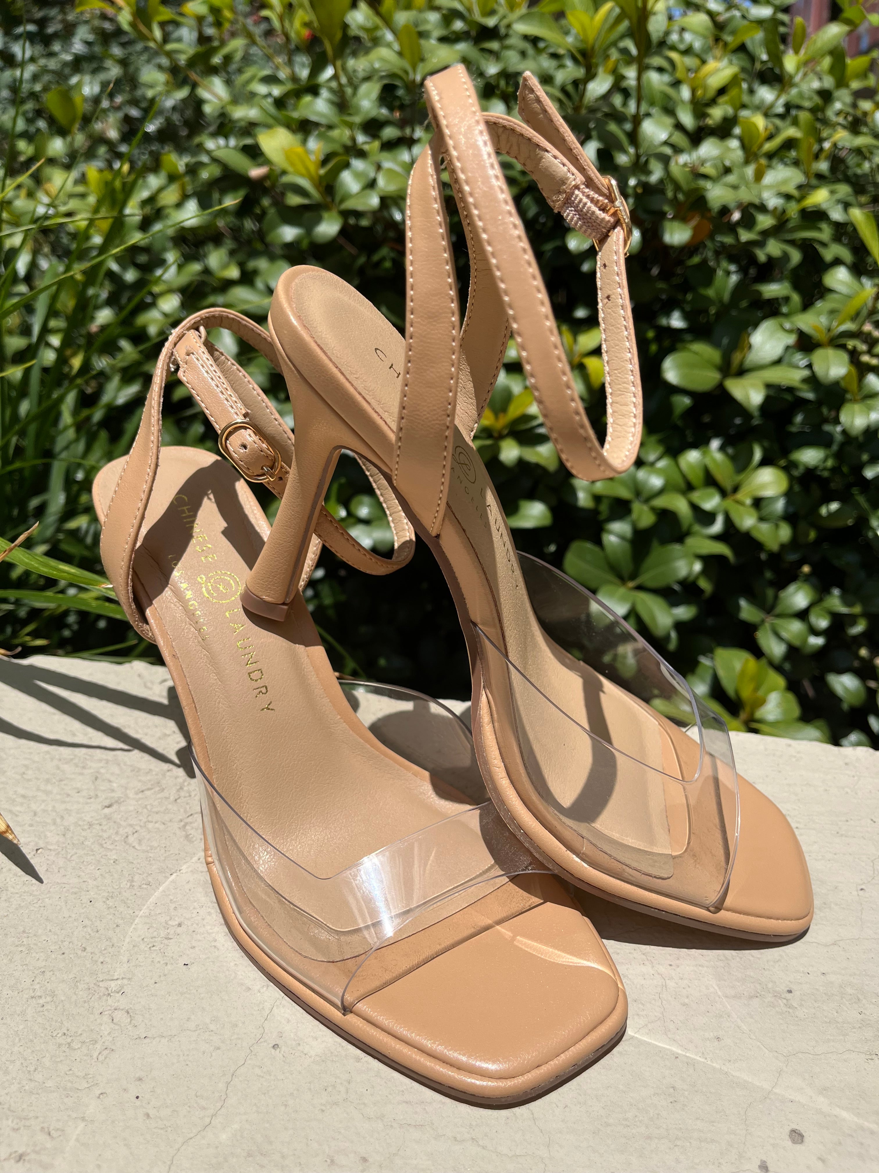 Beige high-heeled sandals with clear straps on a concrete surface with greenery in the background
