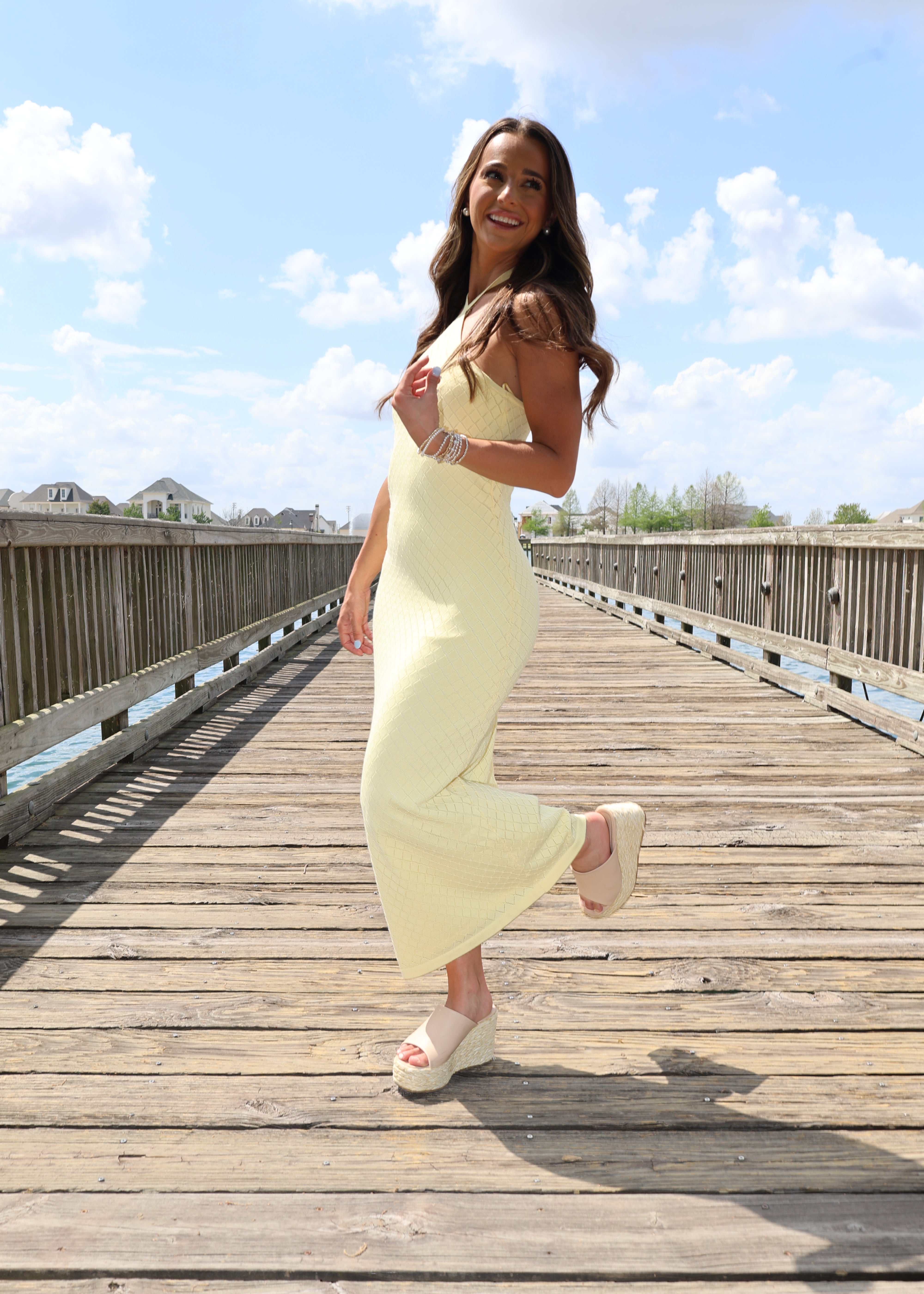 Woman in a yellow dress standing on a wooden boardwalk with a blue sky and clouds in the background.