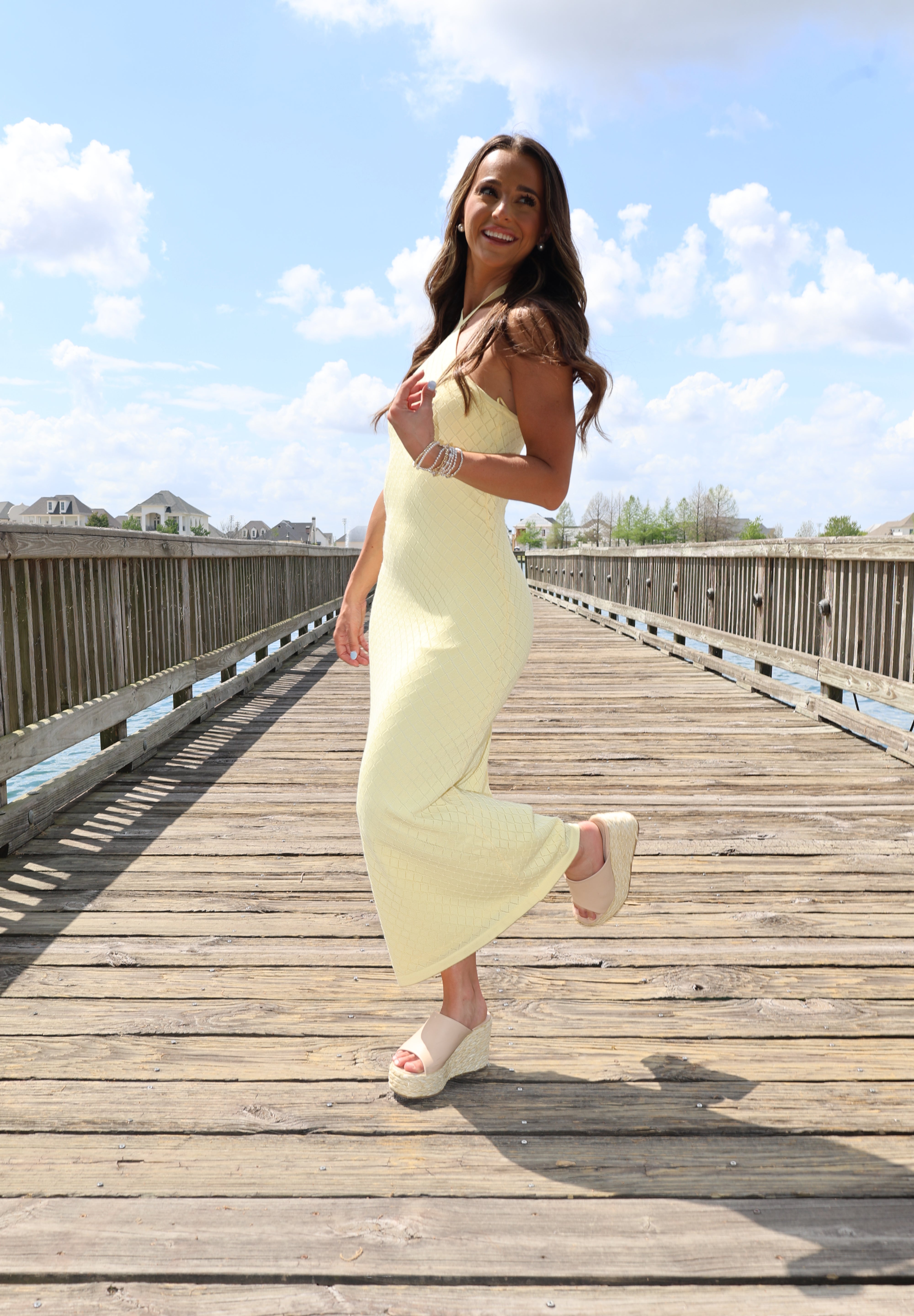 Woman in a yellow dress standing on a wooden boardwalk with a blue sky and clouds in the background.