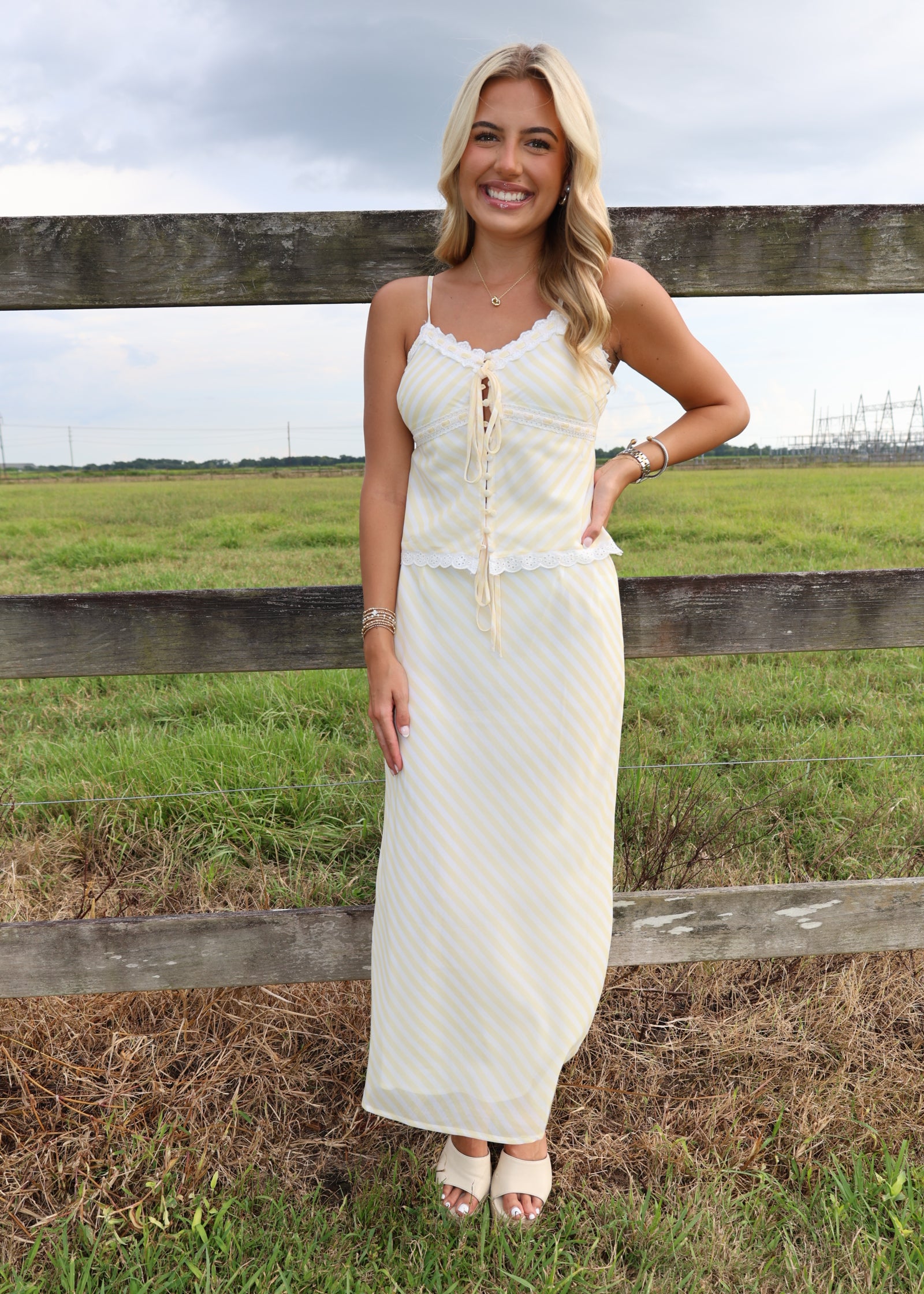 A woman stands outdoors in front of a wooden fence and grassy field, wearing a long cream maxi dress with thin straps, lace-up front, and subtle vertical texture.