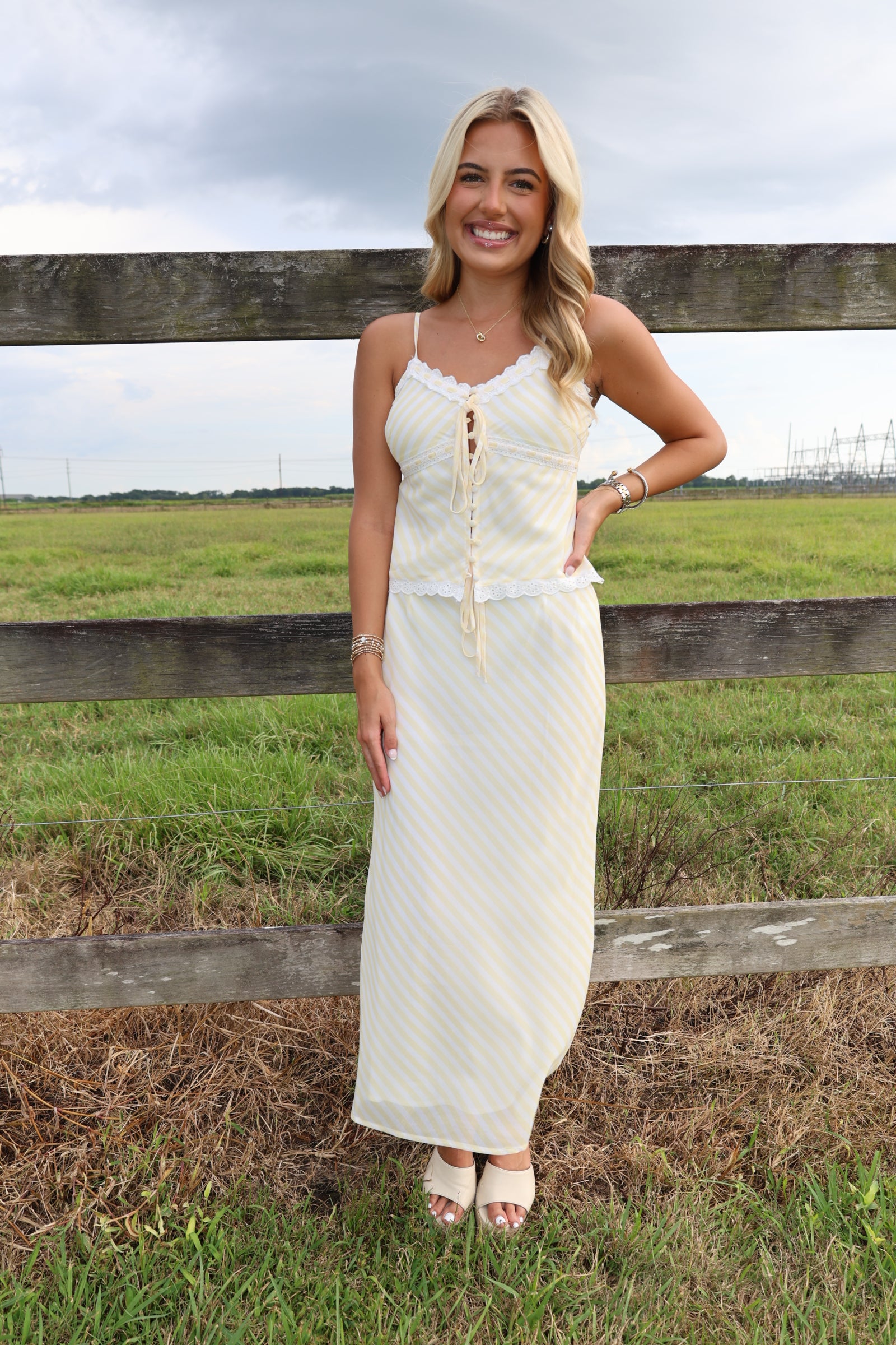 A woman stands outdoors in front of a wooden fence and grassy field, wearing a long cream maxi dress with thin straps, lace-up front, and subtle vertical texture.