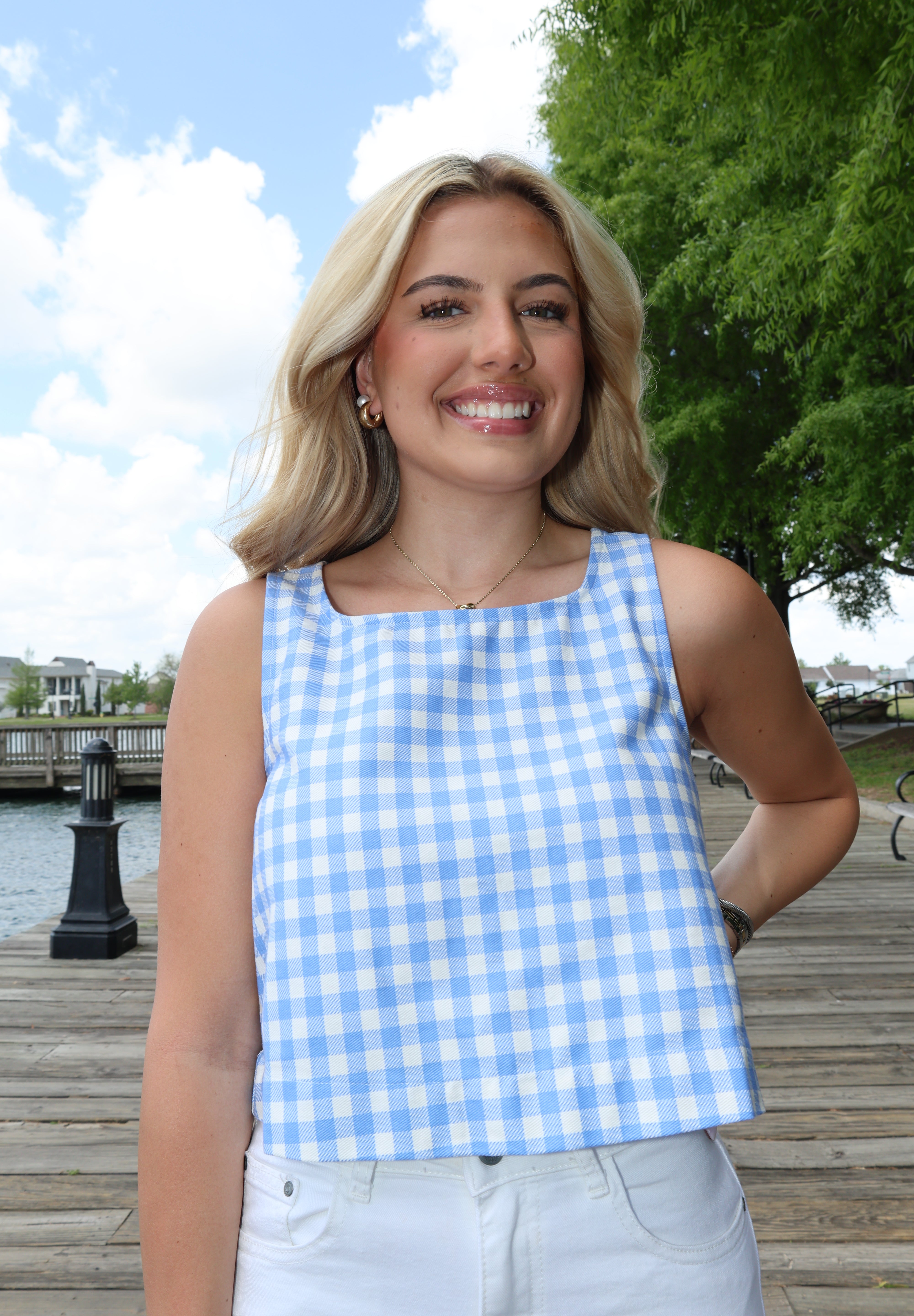 Woman wearing a blue and white checkered dress standing outdoors with trees and a clear sky in the background.