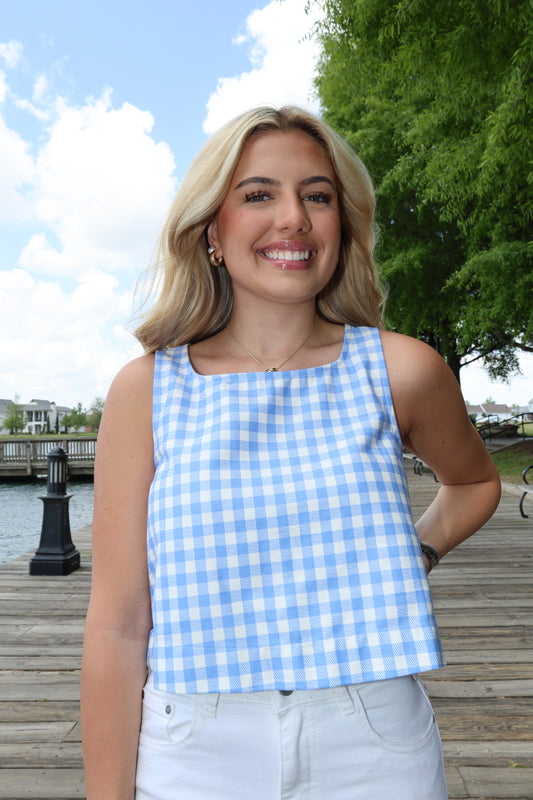 Woman wearing a blue and white checkered dress standing outdoors with trees and a clear sky in the background.
