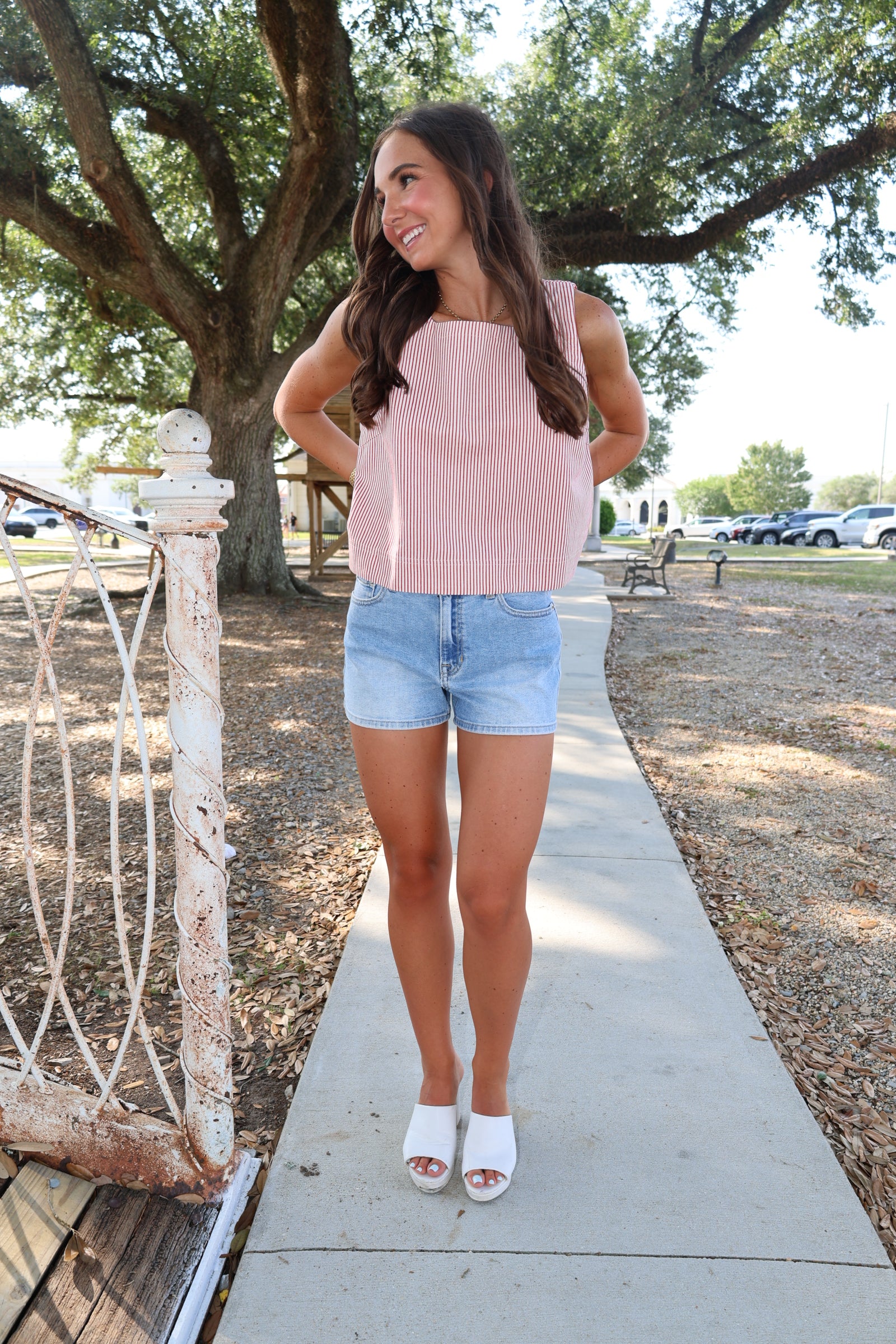 Woman in a pink top and denim shorts standing on a sidewalk with trees in the background
