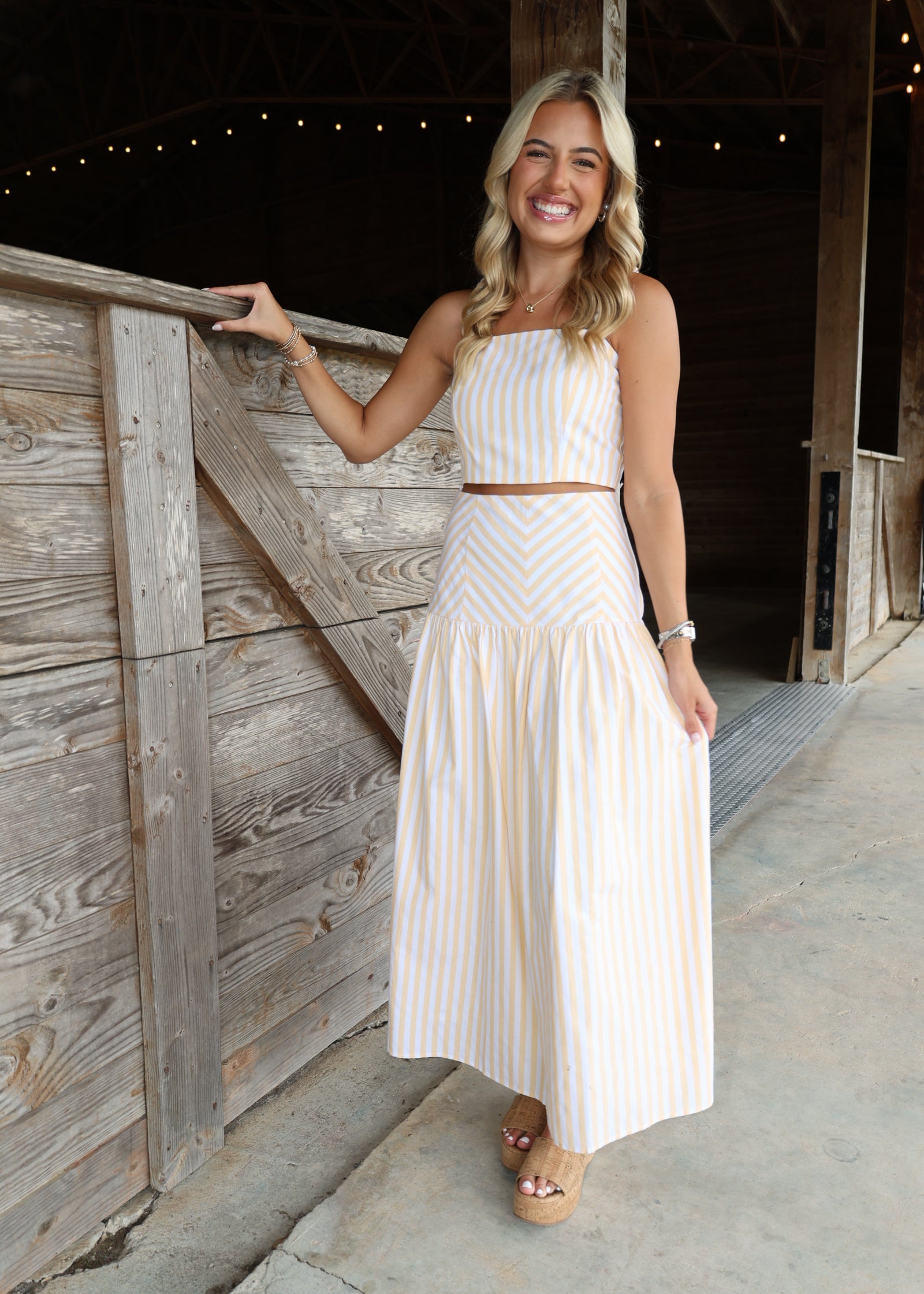 A woman stands in a rustic barn doorway wearing a sleeveless, two-piece cream and light pink striped dress with a halter top and a long pleated skirt.