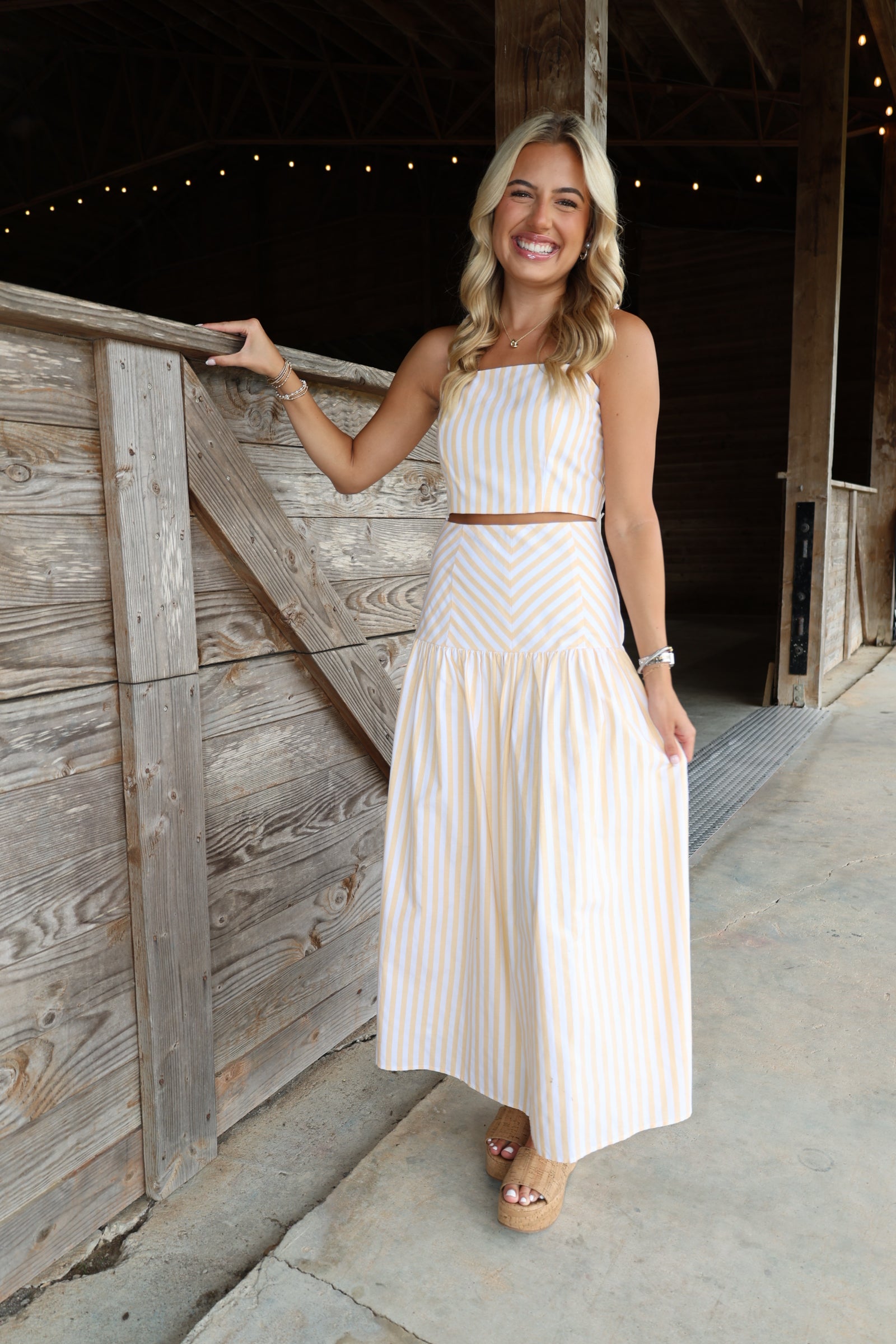 A woman stands in a rustic barn doorway wearing a sleeveless, two-piece cream and light pink striped dress with a halter top and a long pleated skirt.