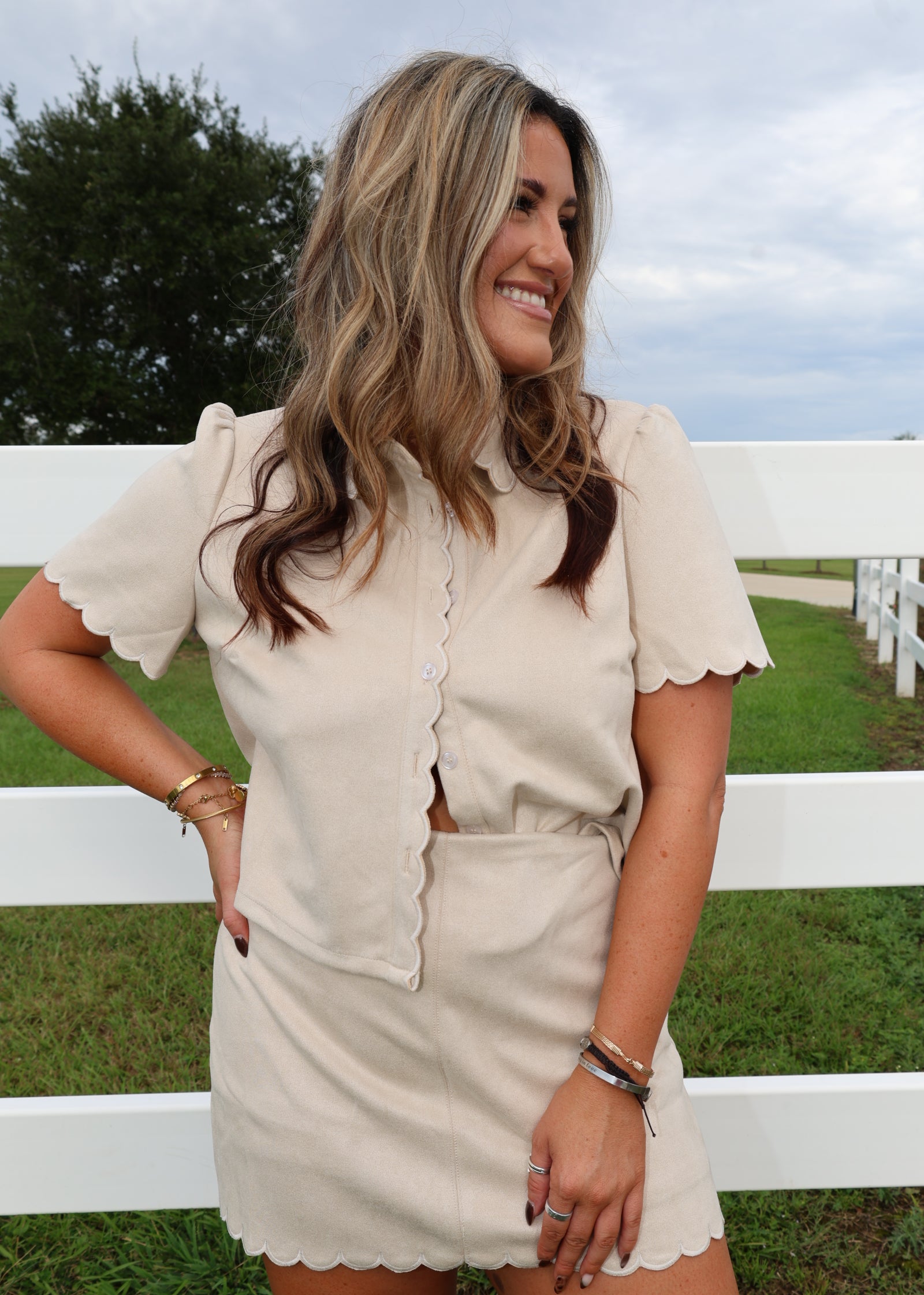 A woman wearing a cream-colored short-sleeve button-up shirt and matching scalloped mini skirt, posing outdoors in front of a white fence.