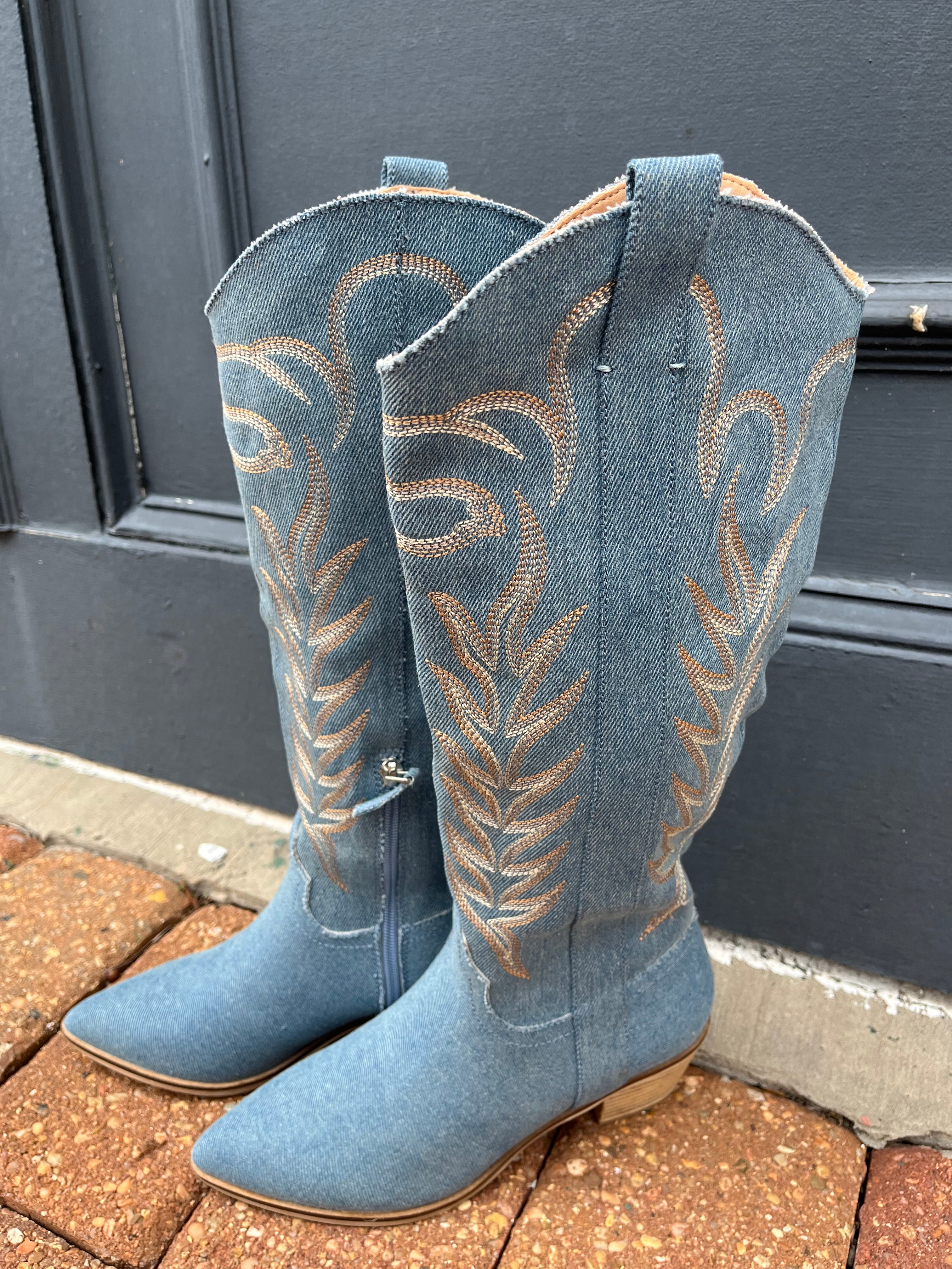 A pair of blue denim boots with brown stitching and pointed toes, displayed against a door.