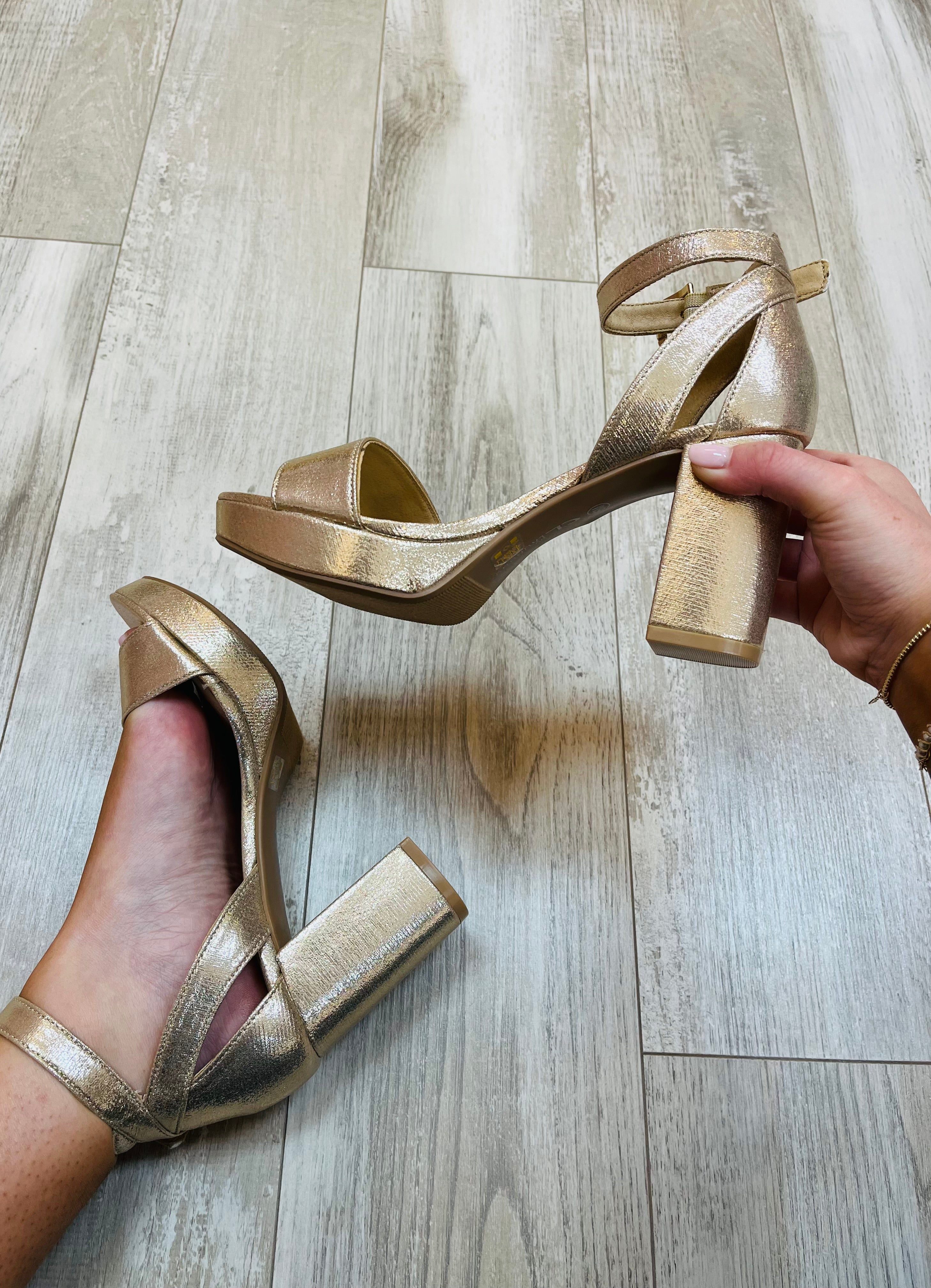 A pair of gold strappy sandals with heels, held up against a light background.