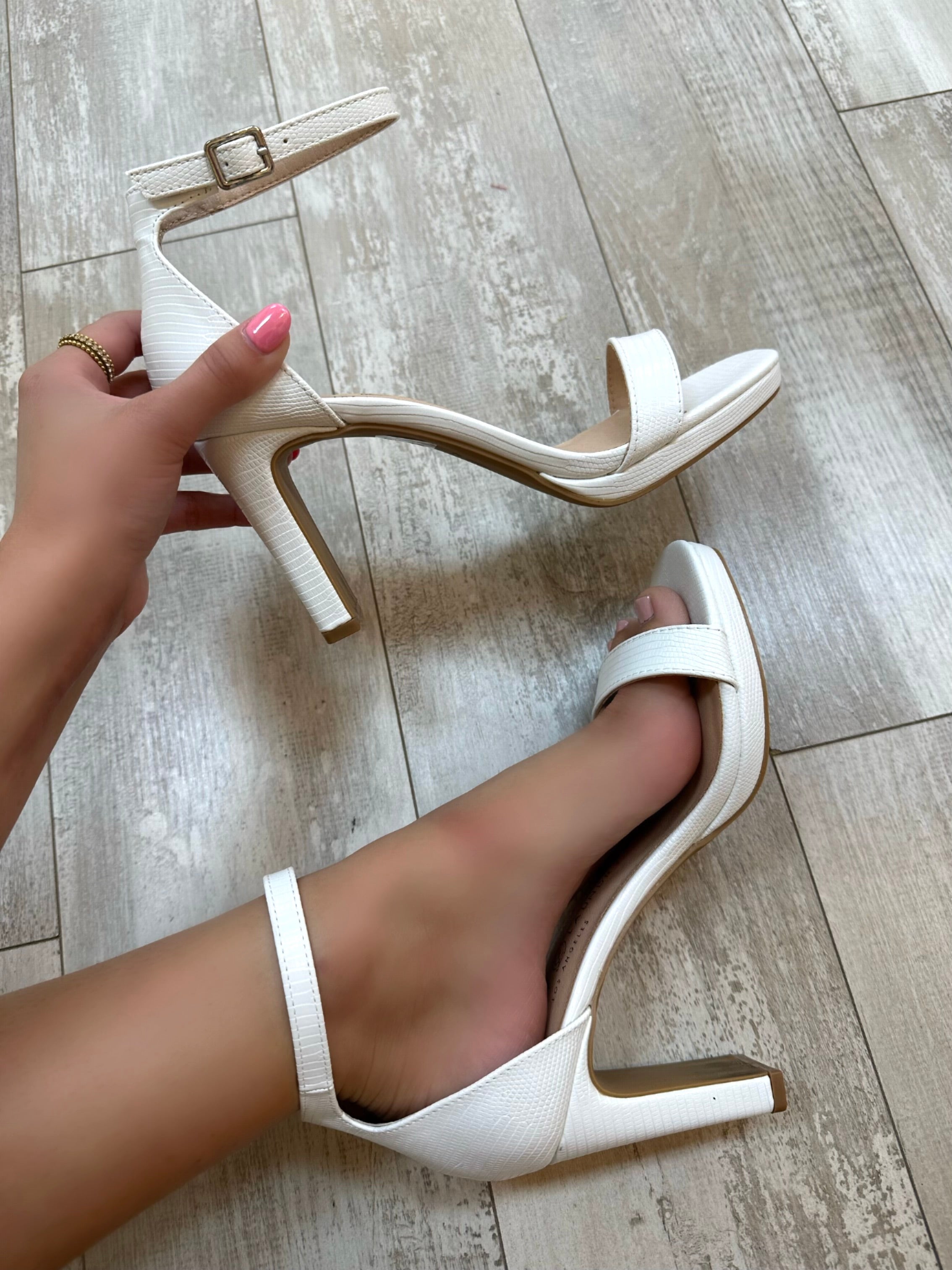 A person standing on a tiled floor is wearing white lizard patterned open-toe platform heels with ankle straps.