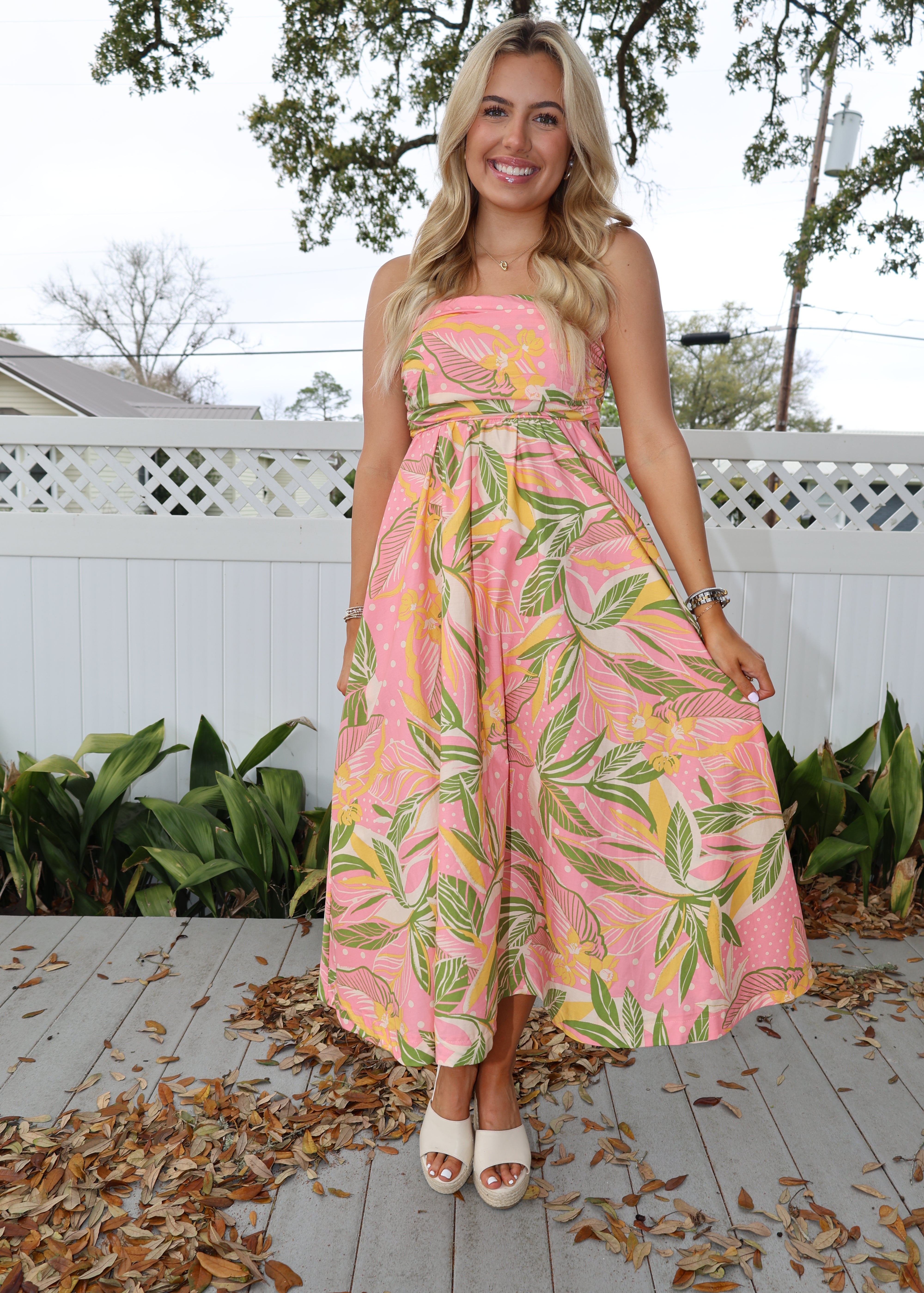 Woman wearing a pink floral dress standing on a wooden deck with plants and trees in the background.