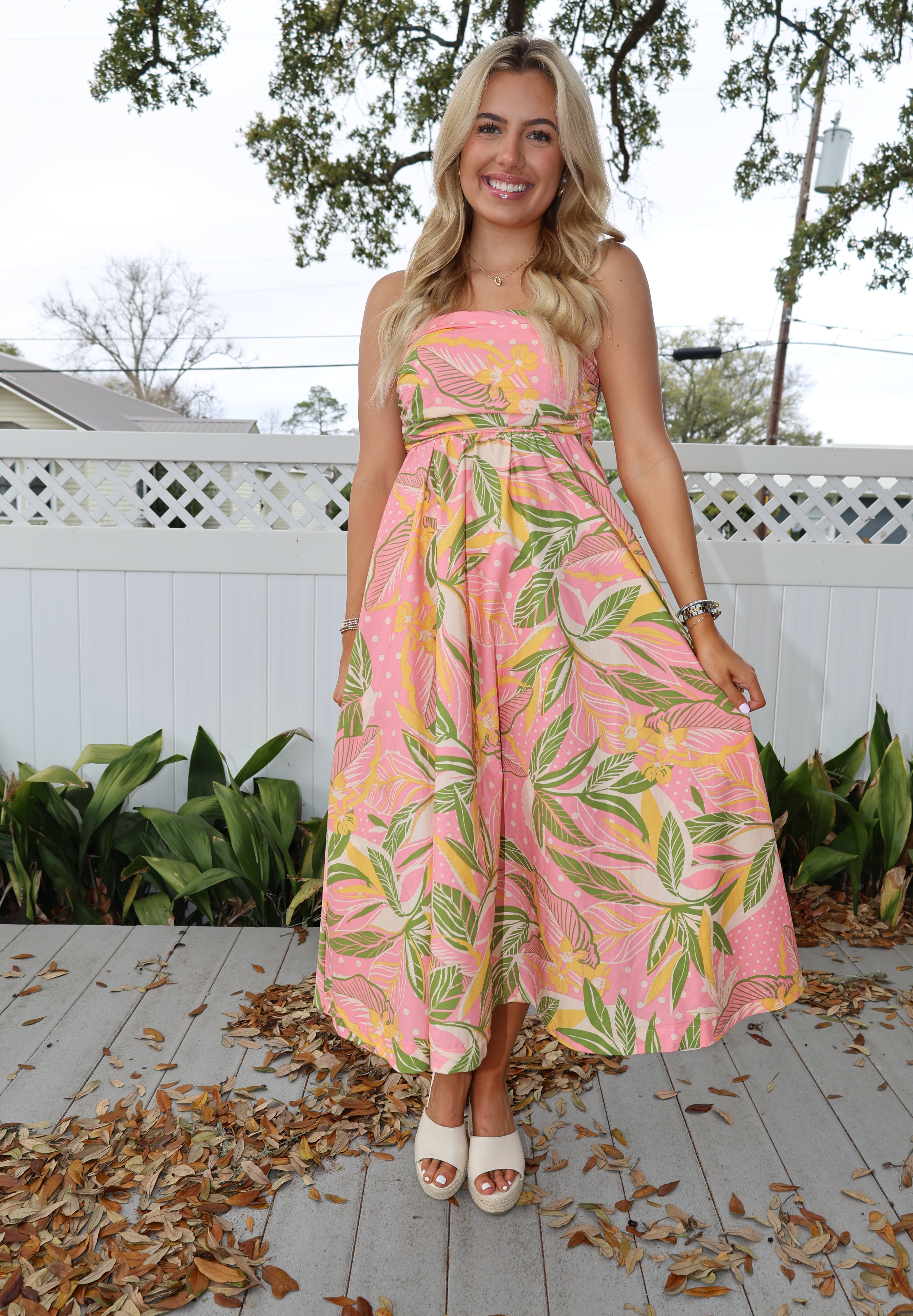 Woman wearing a pink floral dress standing on a wooden deck with plants and trees in the background.