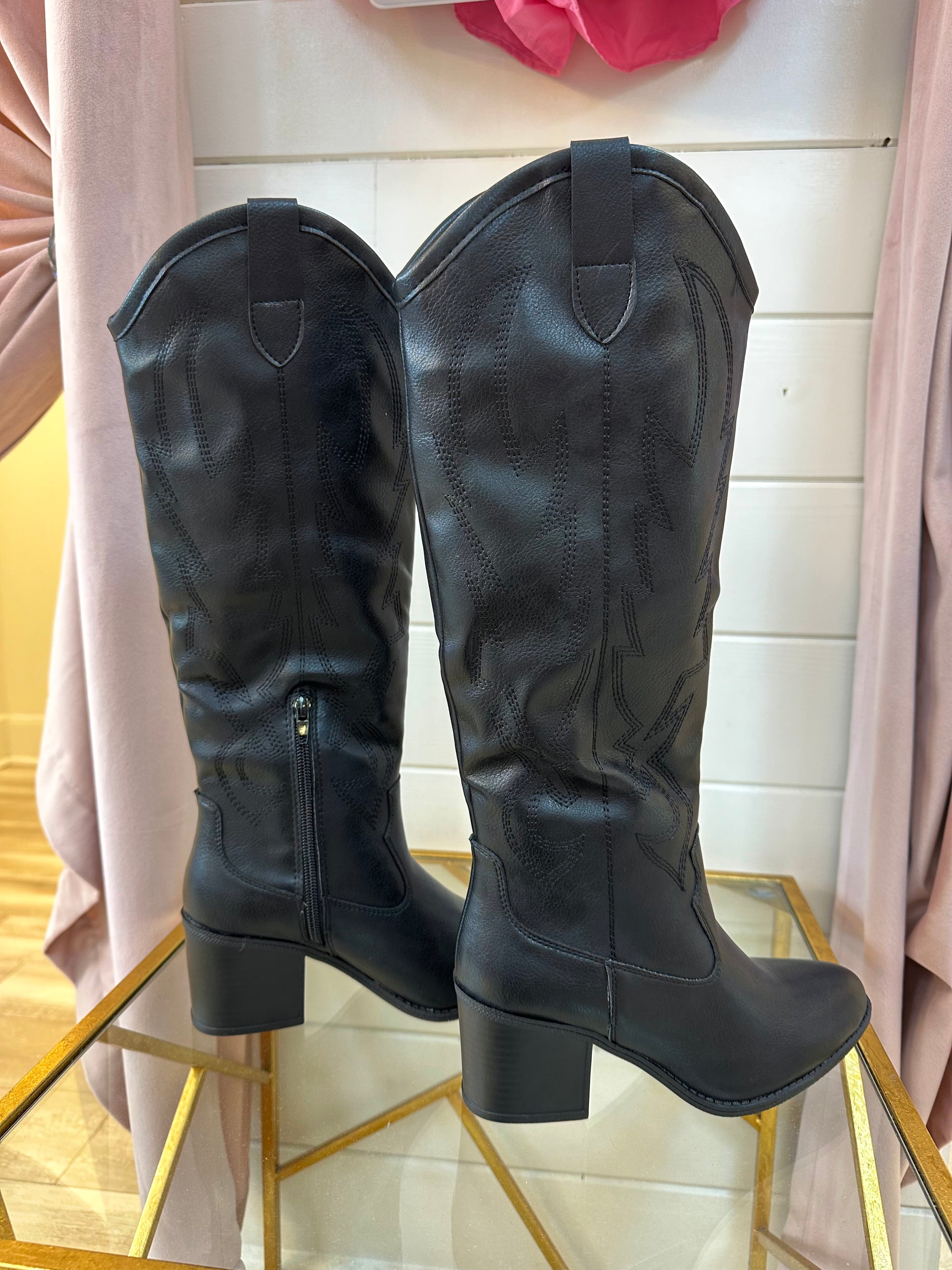 A pair of black, tall boots displayed on a shoe stand with a curtain and pink flower in the background.