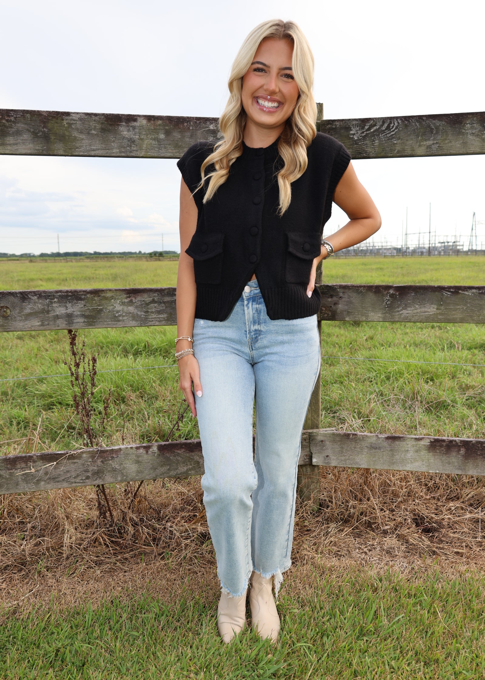 A woman stands outdoors in front of a wooden fence and grassy field, wearing a sleeveless black button-front top, light-wash blue jeans, and beige ankle boots.