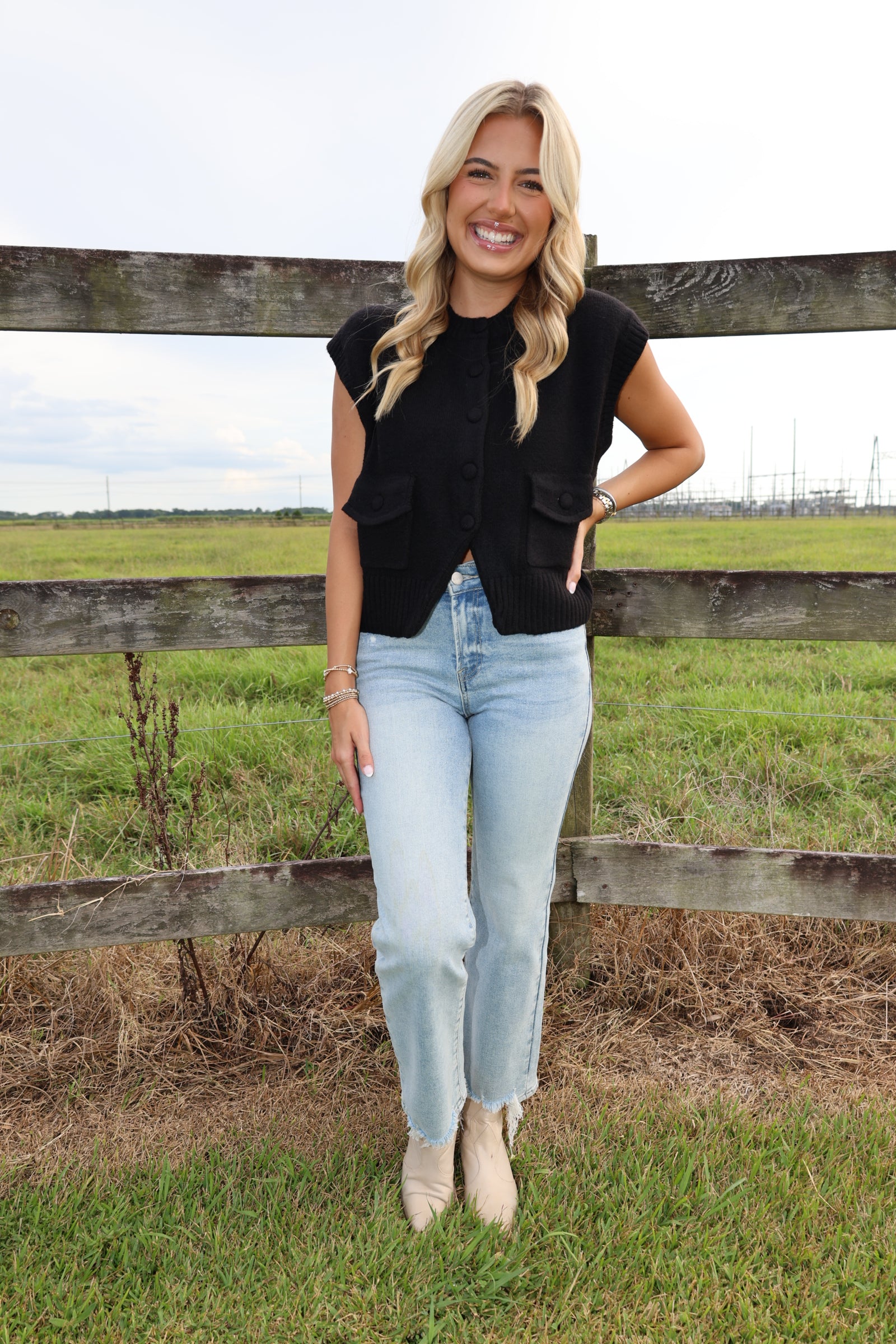 A woman stands outdoors in front of a wooden fence and grassy field, wearing a sleeveless black button-front top, light-wash blue jeans, and beige ankle boots.