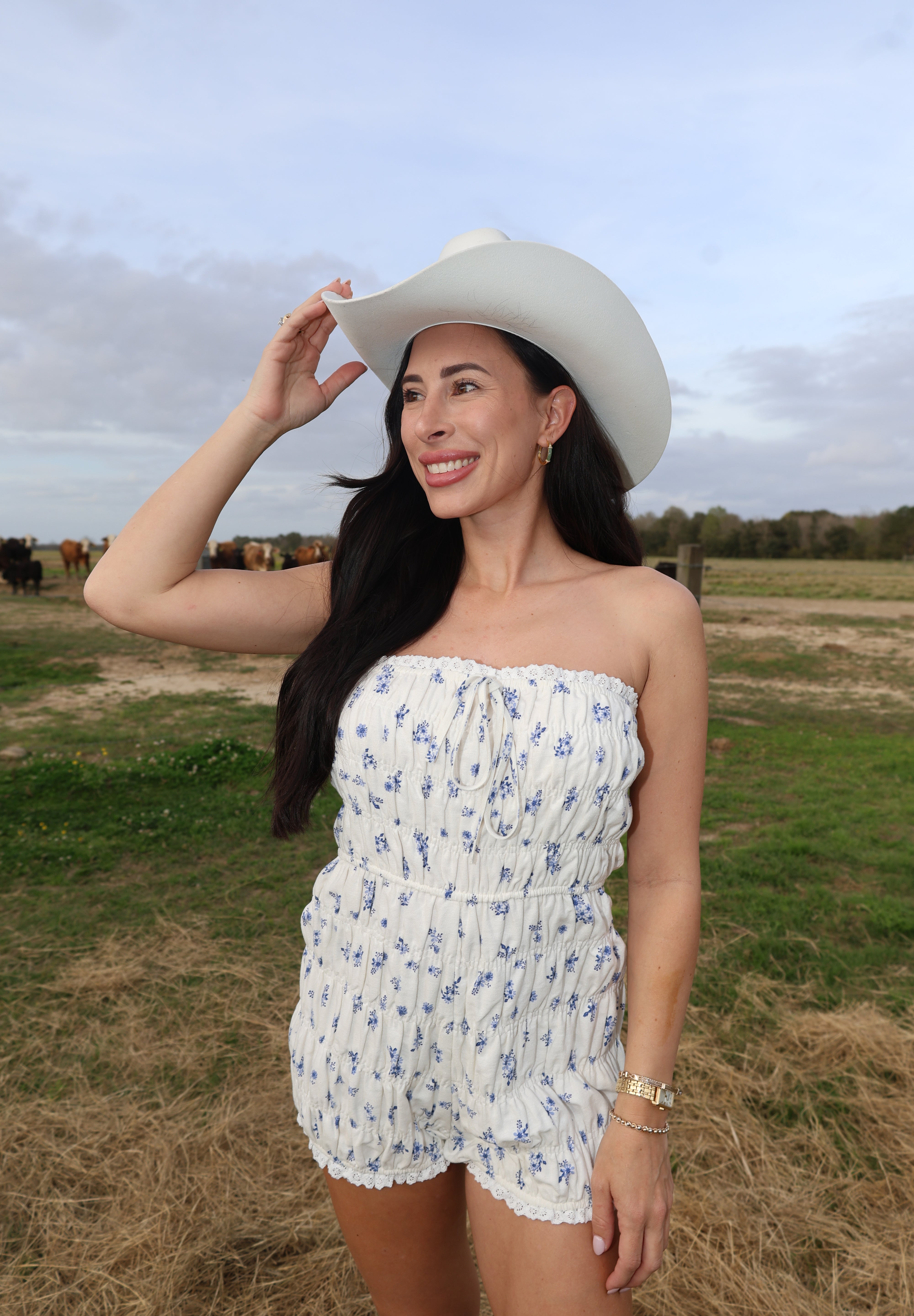 Woman in a white dress with blue patterns and a white hat standing in a field with water in the background.