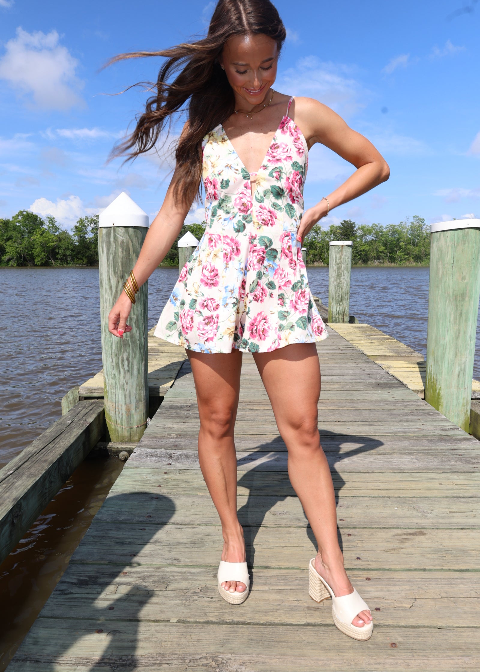 Woman in a floral dress standing on a wooden dock by a body of water.