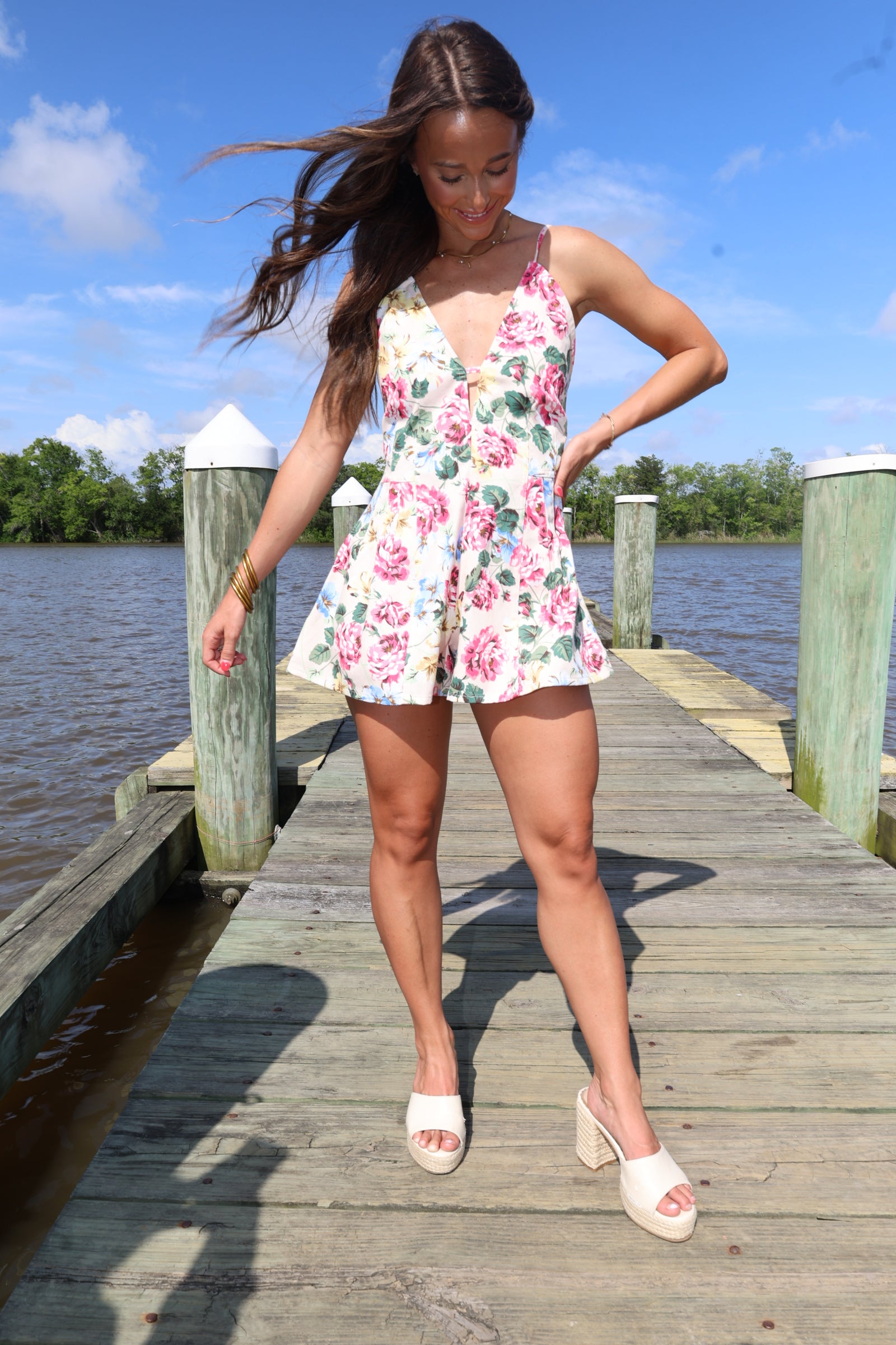 Woman in a floral dress standing on a wooden dock by a body of water.