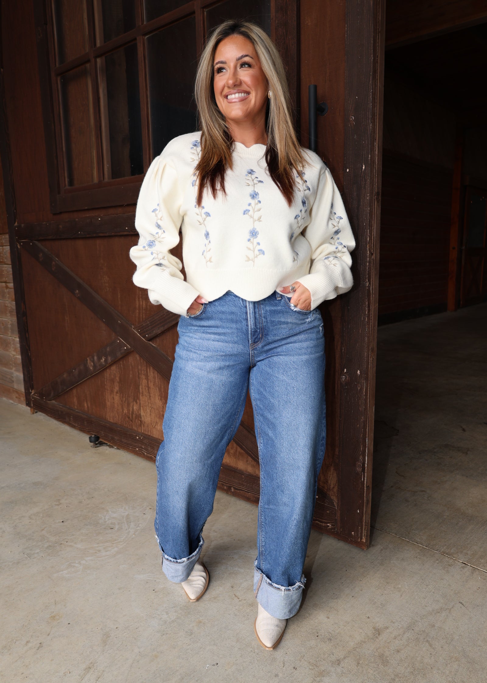 A woman stands in front of a rustic wooden barn door wearing a cream-colored long-sleeve sweater with blue floral embroidery, blue jeans, and beige pointed-toe heels.