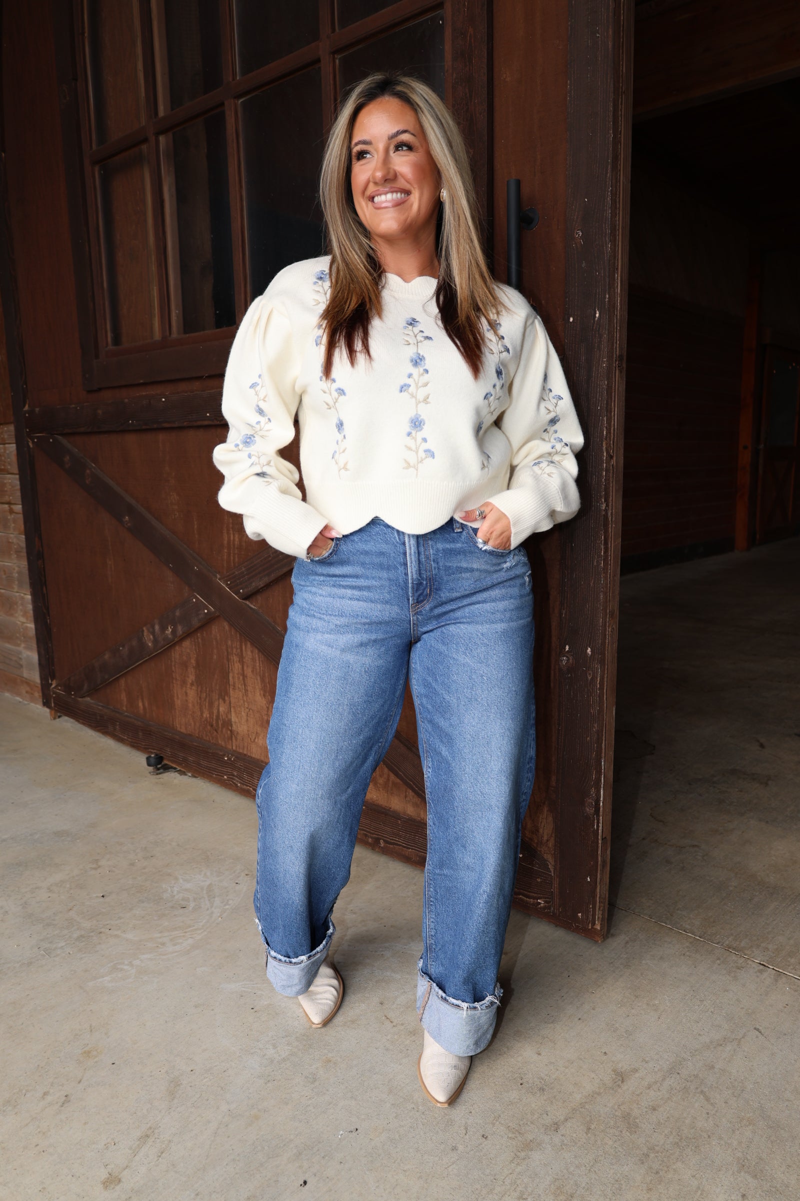 A woman stands in front of a rustic wooden barn door wearing a cream-colored long-sleeve sweater with blue floral embroidery, blue jeans, and beige pointed-toe heels.