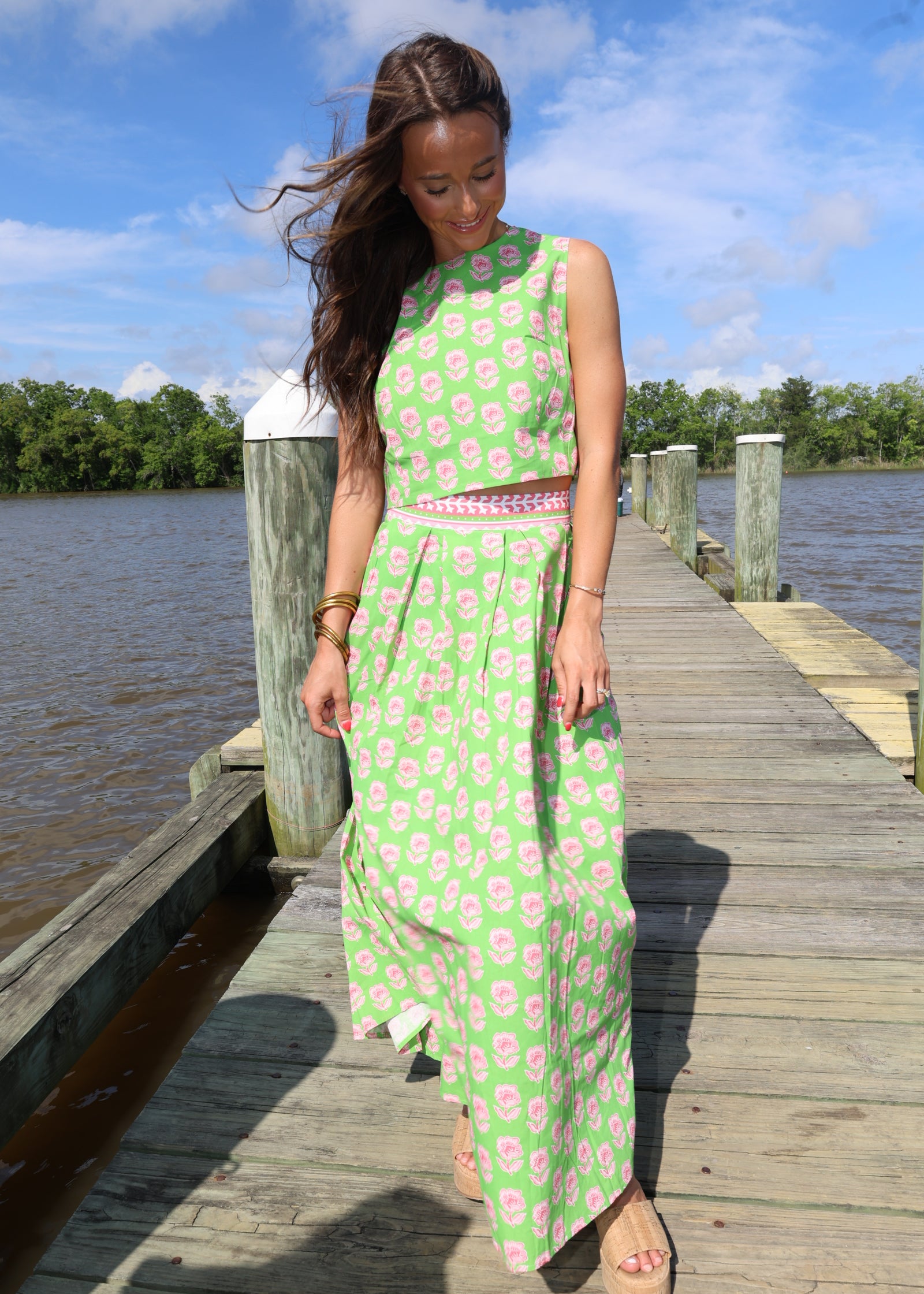 Woman in a green dress with pink floral pattern standing on a wooden dock by water.