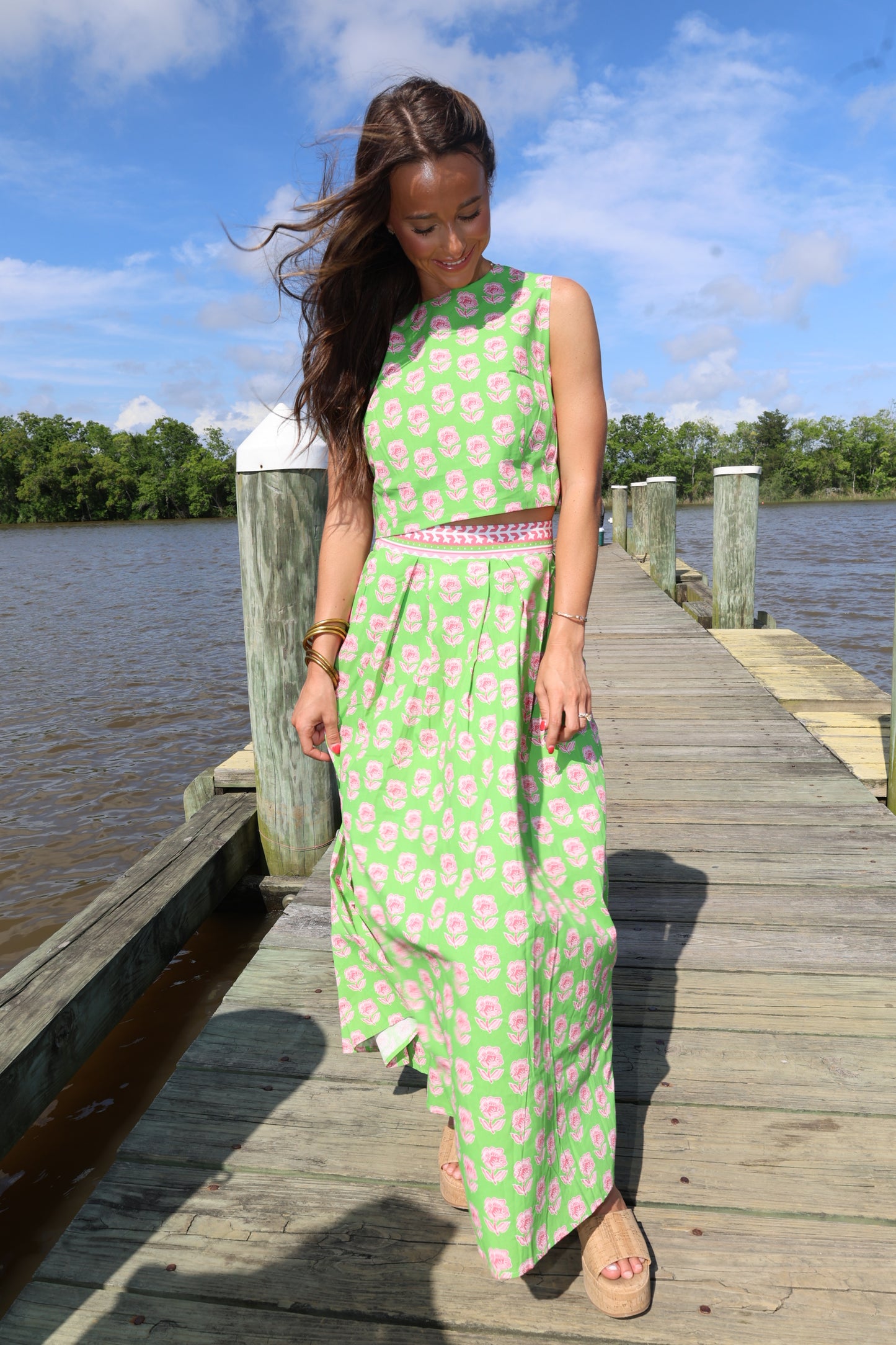 Woman in a green dress with pink floral pattern standing on a wooden dock by water.