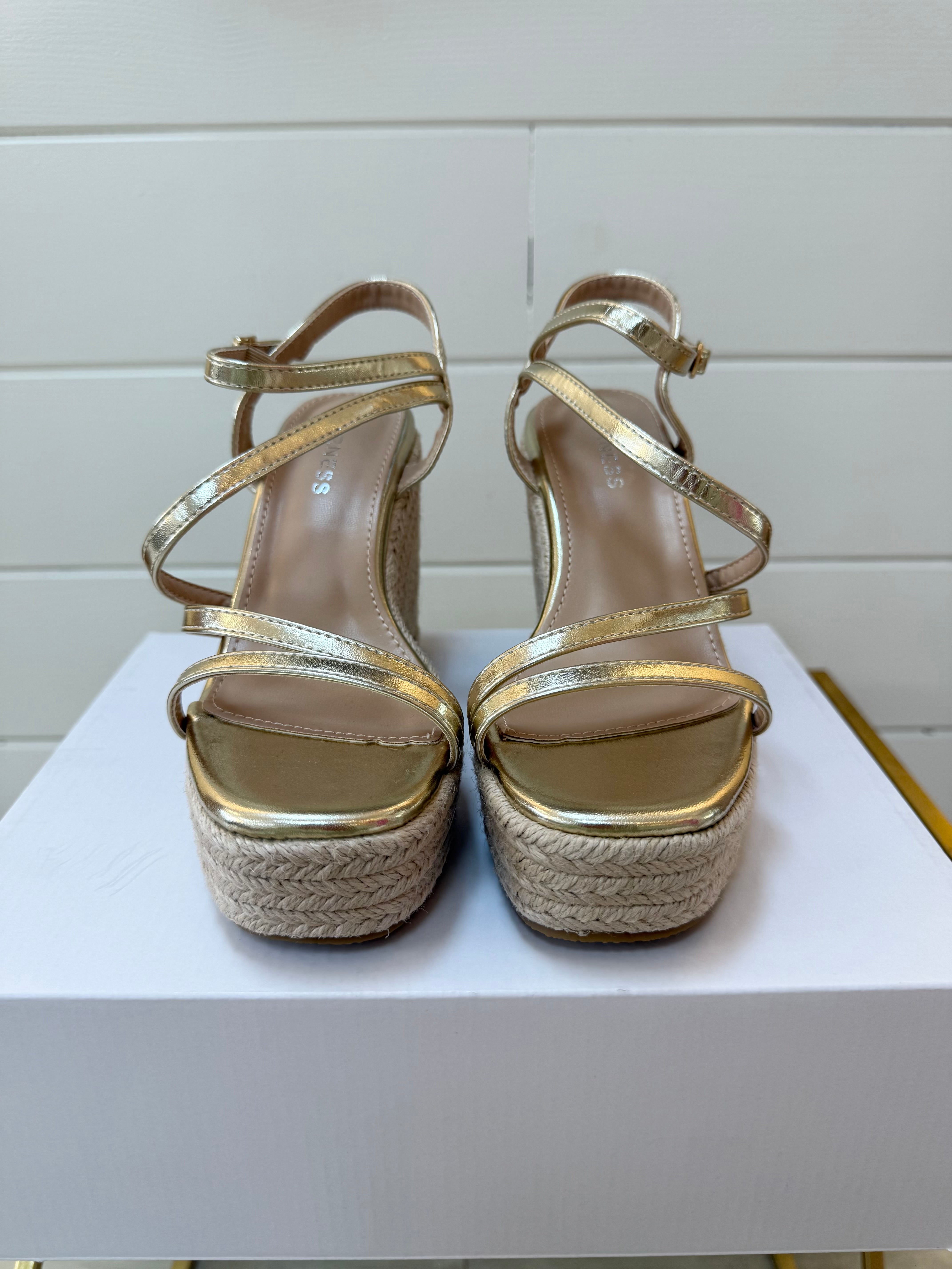 Gold espadrille wedge sandals on a white surface with a light wooden floor background.