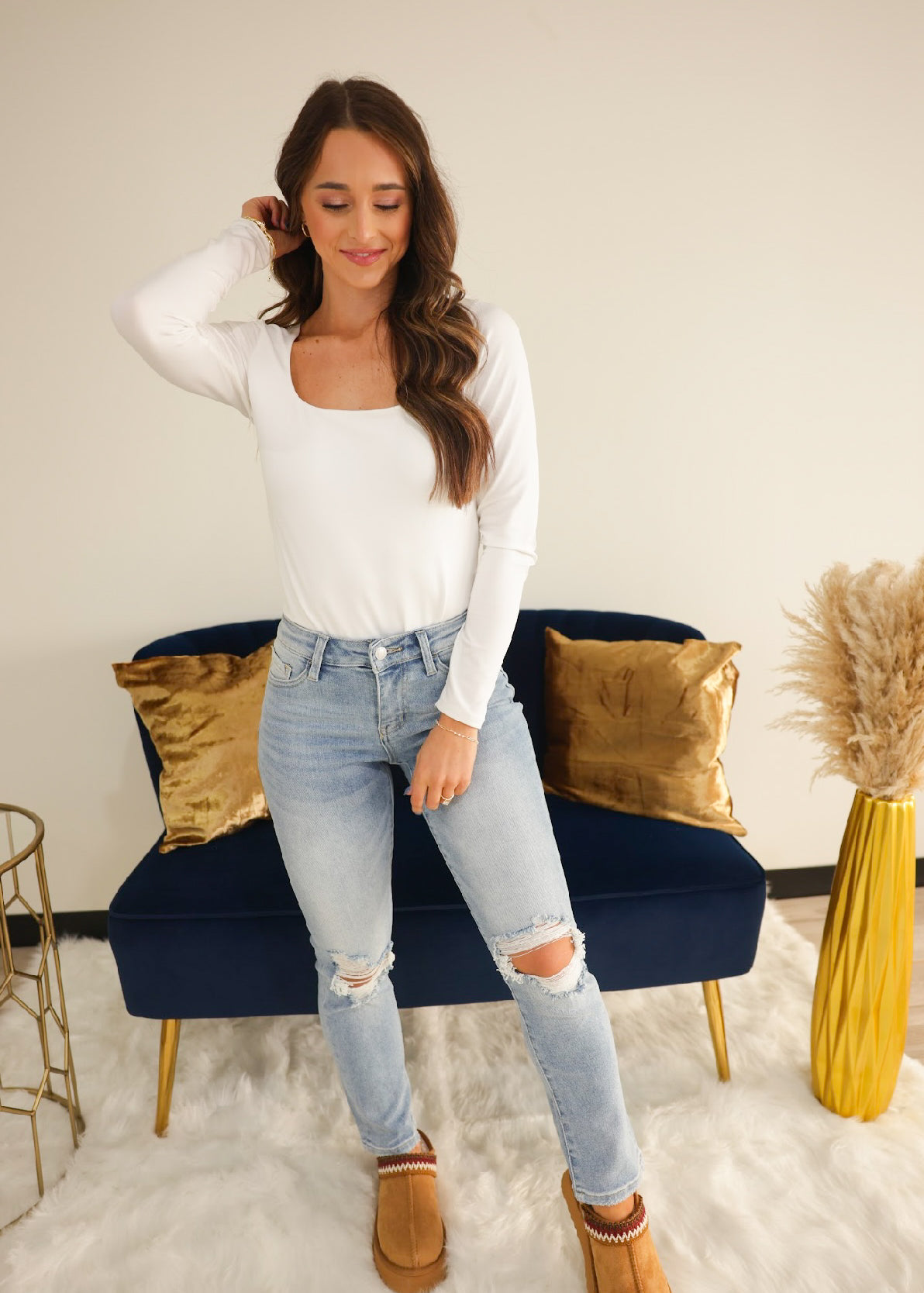 A woman standing in a room with blue denim jeans, a white long-sleeve top, and tan shoes.