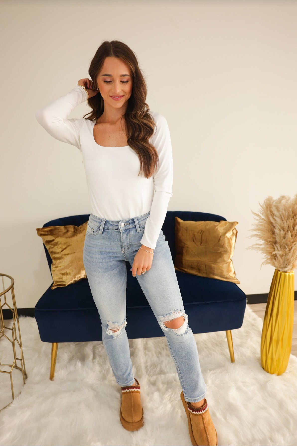 A woman standing in a room with blue denim jeans, a white long-sleeve top, and tan shoes.