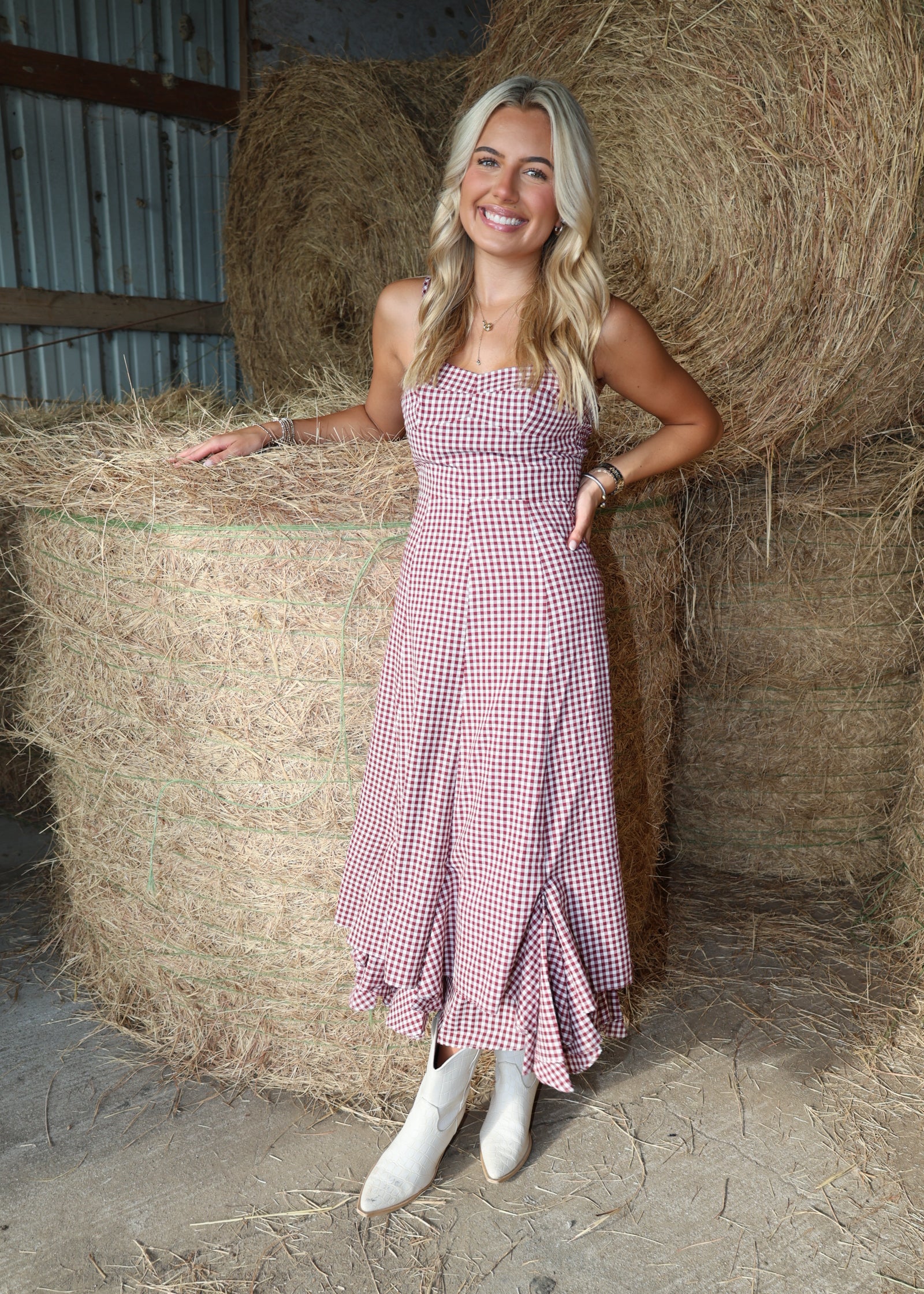 A woman stands in a barn in front of large hay bales, wearing a pink and white gingham strapless midi dress with a ruffled hem and white cowboy boots.