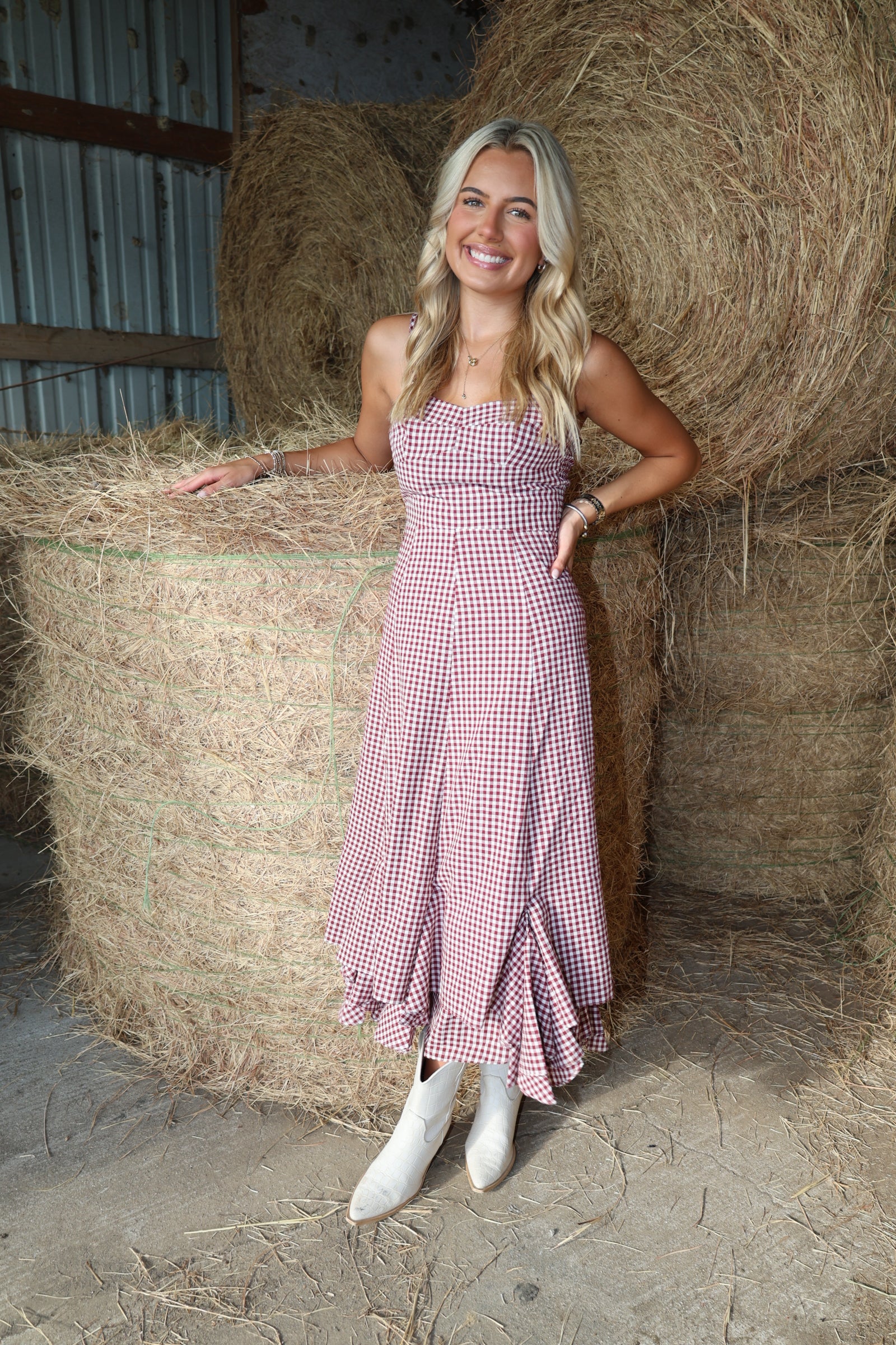 A woman stands in a barn in front of large hay bales, wearing a pink and white gingham strapless midi dress with a ruffled hem and white cowboy boots.