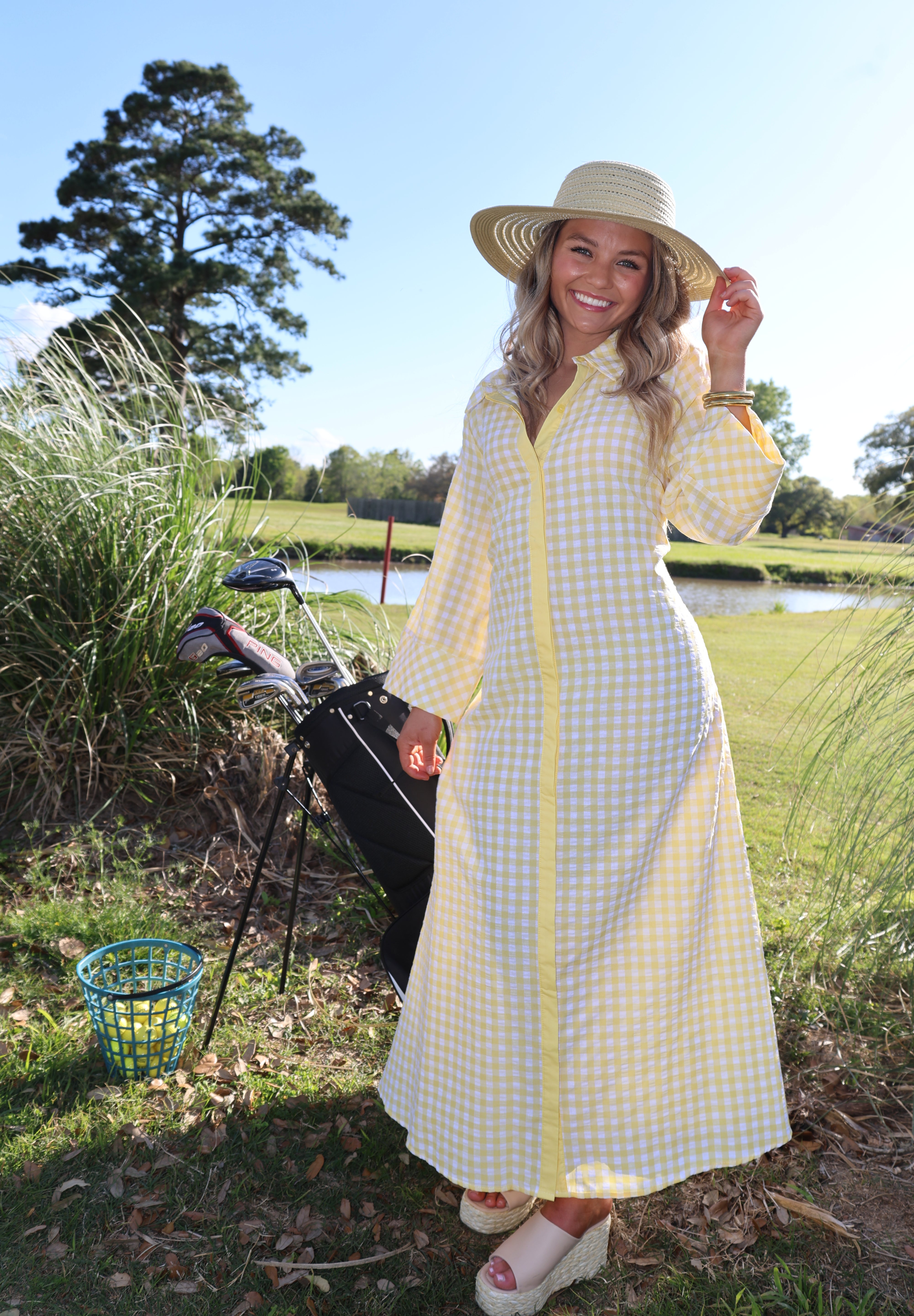 Woman in a yellow checkered dress and hat standing outdoors with golf equipment.