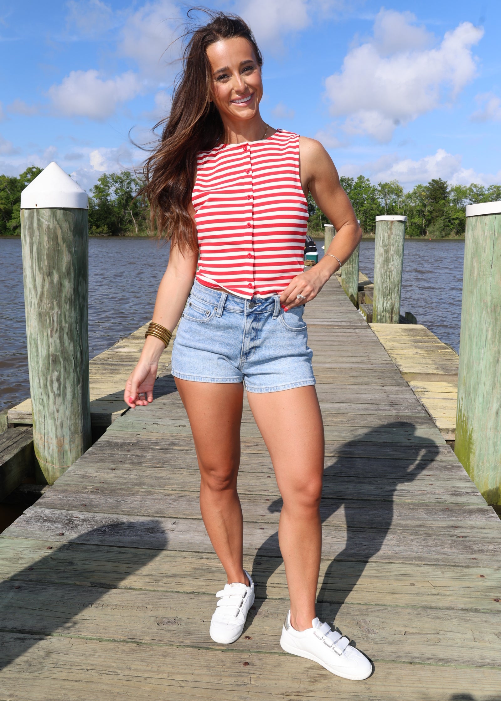 Woman standing on a wooden dock by a body of water with a blue sky and clouds in the background.