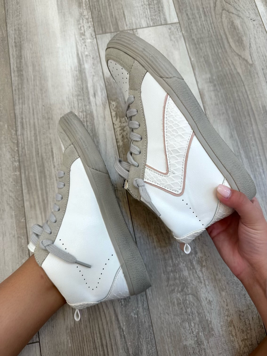 A pair of high-top sneakers with white uppers, grey accents, and beige laces, held by a person against a wooden background.