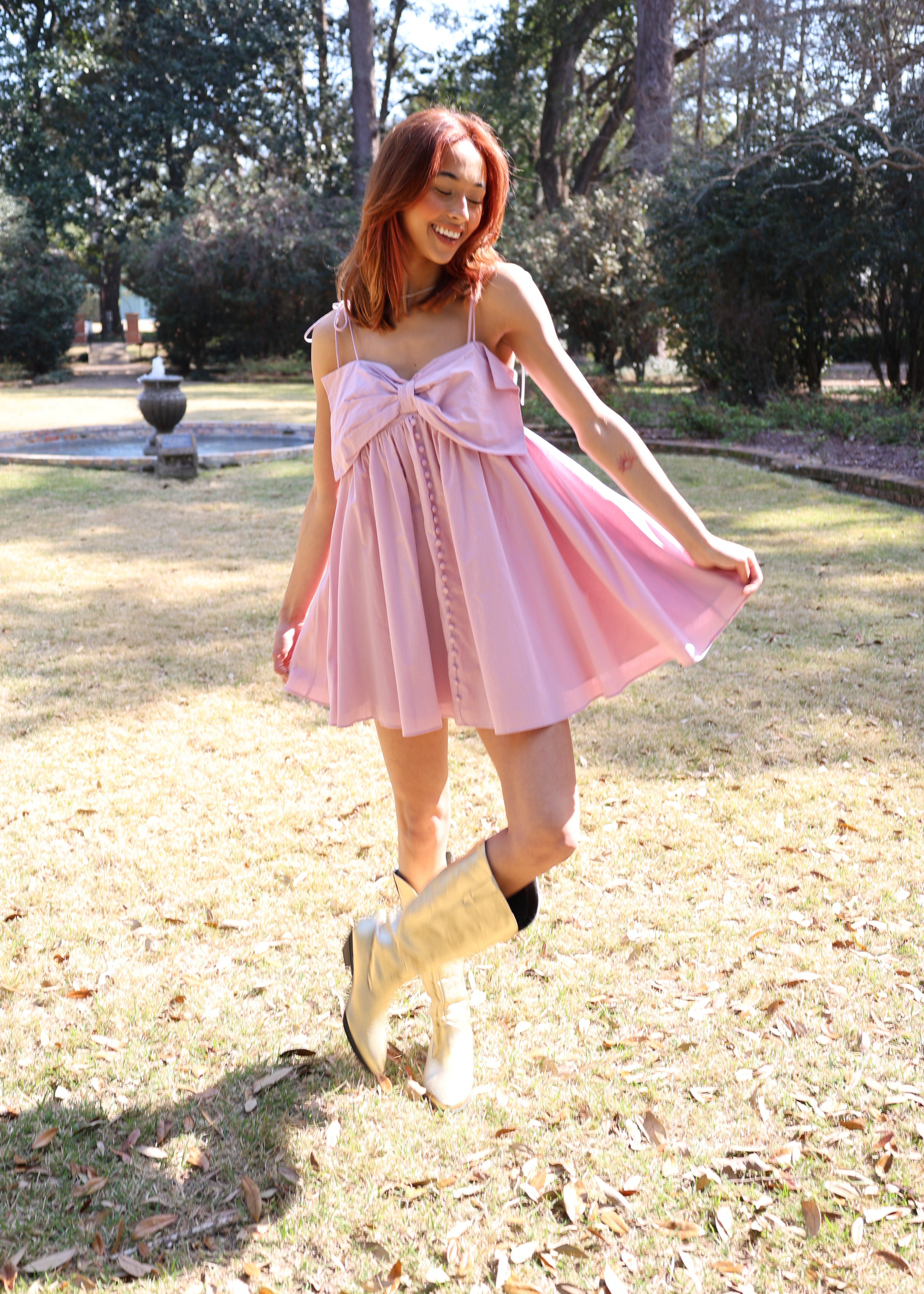 A person wearing a lavender dress with a pleated skirt and bow detail at the chest, paired with yellow boots, standing in an outdoor setting.