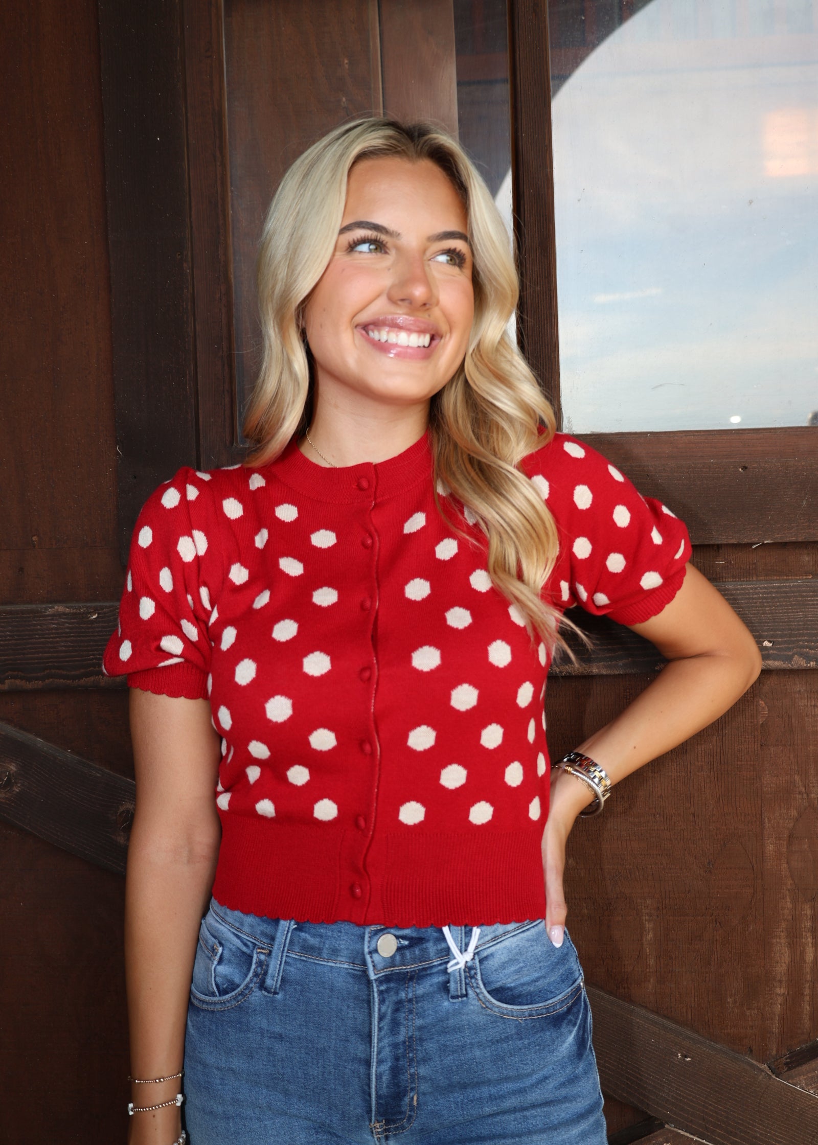 A smiling blonde woman wearing a red short-sleeve button-front cardigan with white polka dots and blue jeans, standing in front of a dark wooden door.
