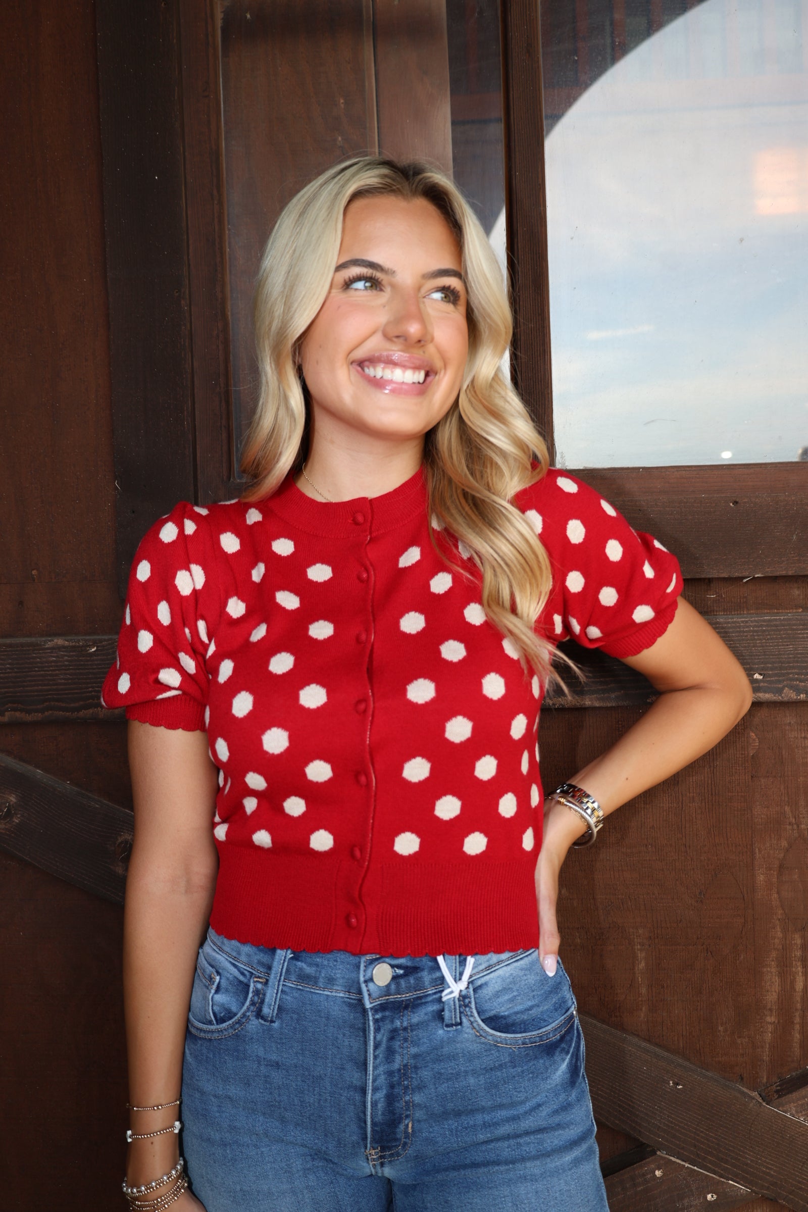 A smiling blonde woman wearing a red short-sleeve button-front cardigan with white polka dots and blue jeans, standing in front of a dark wooden door.