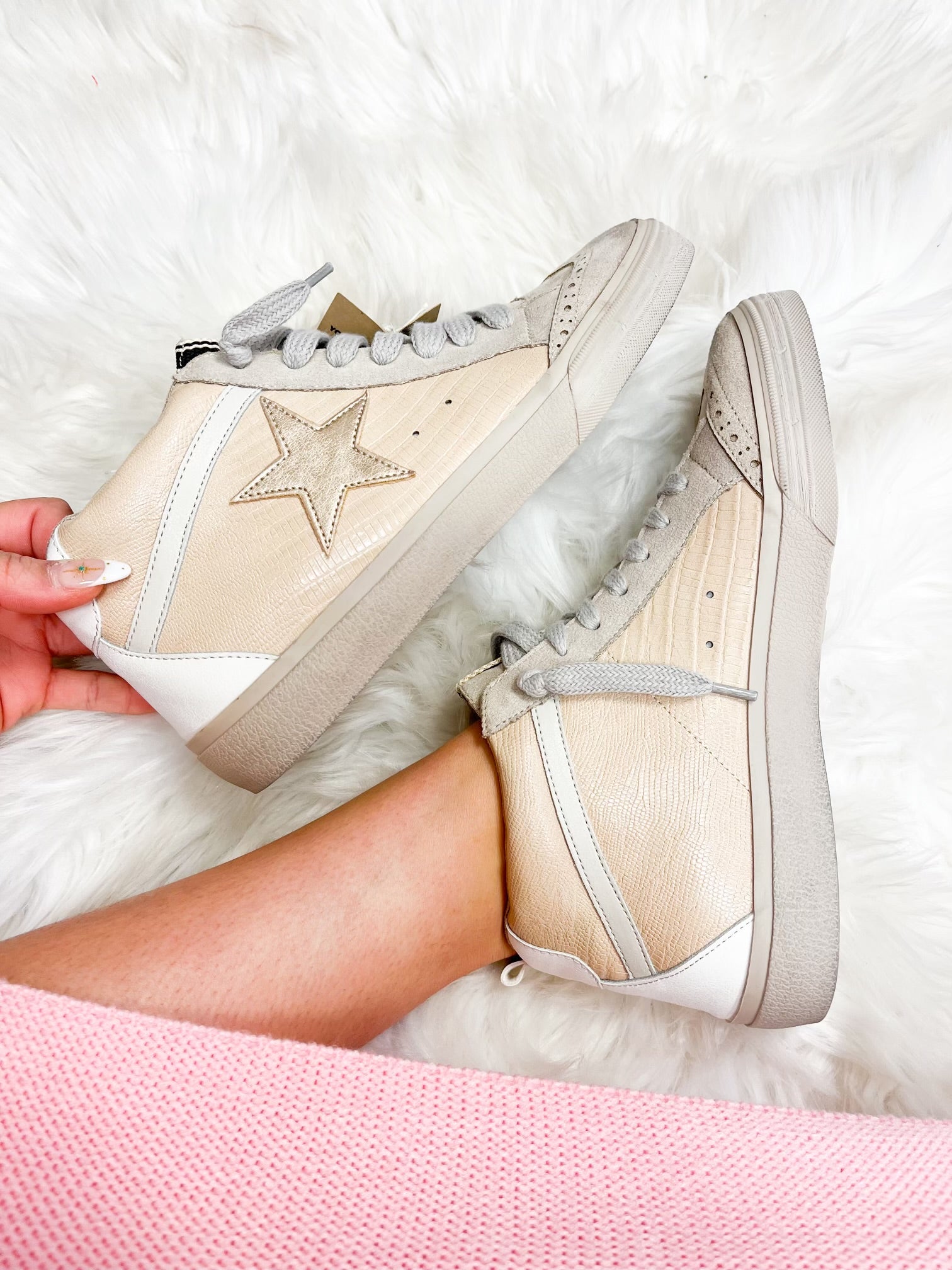 A pair of beige high-top sneakers with a star embellishment on the side, displayed on a person's feet against a white furry background.