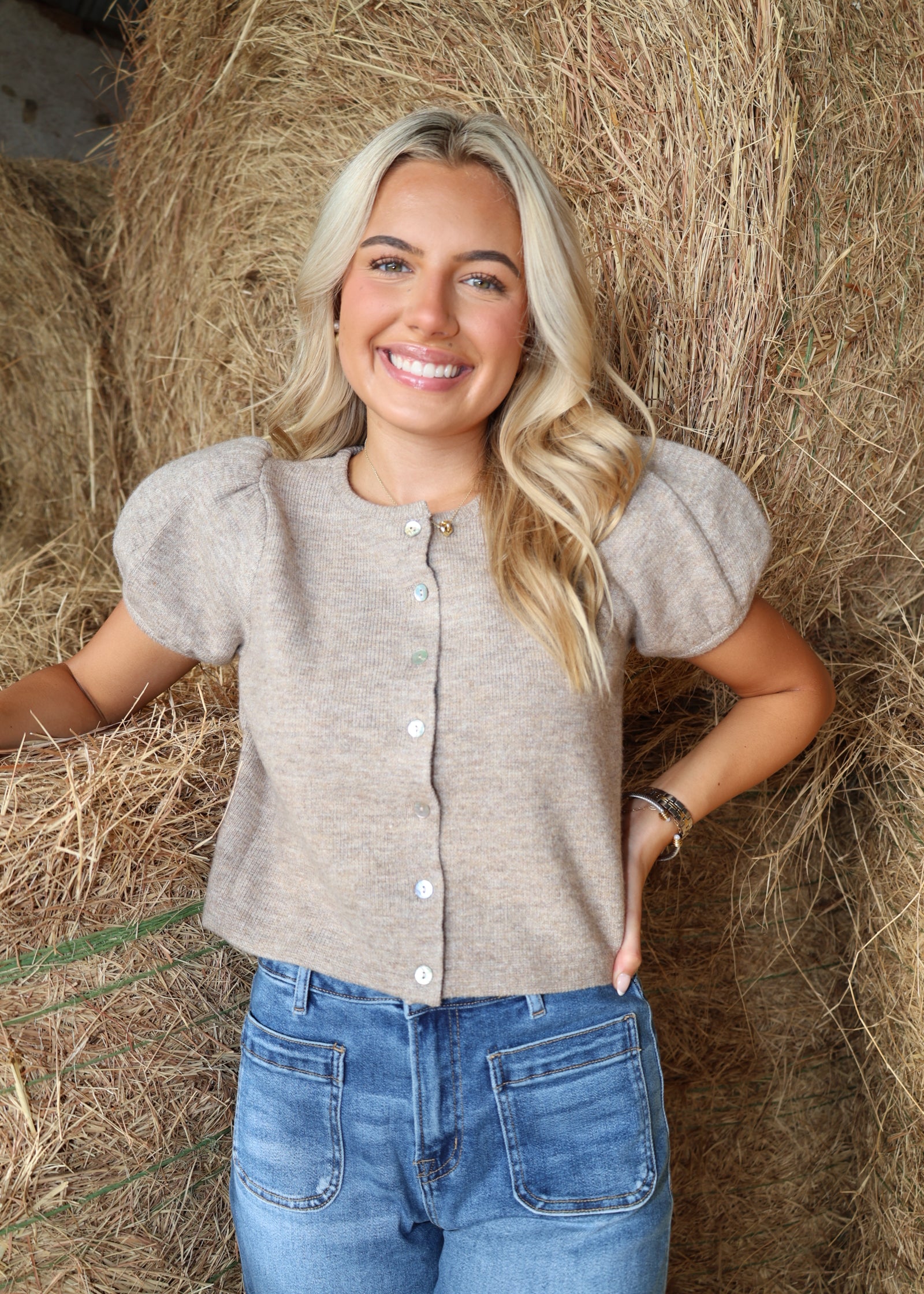 A smiling blonde woman stands in front of a large hay bale wearing a light taupe short-sleeve knit top with puff sleeves and white buttons, paired with blue jeans.