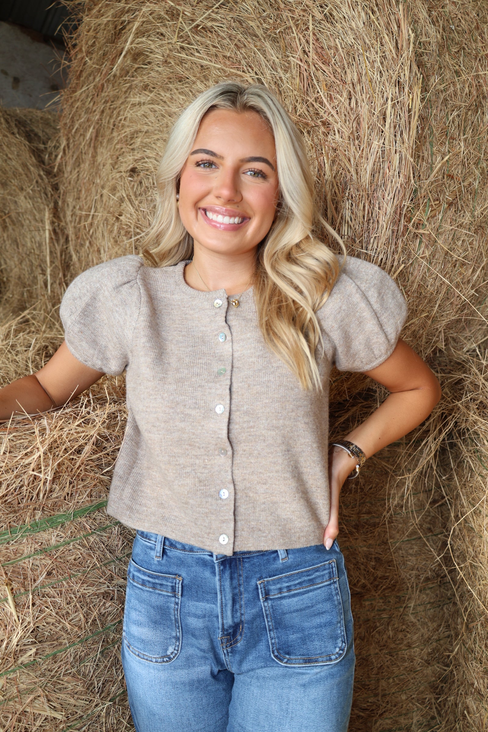 A smiling blonde woman stands in front of a large hay bale wearing a light taupe short-sleeve knit top with puff sleeves and white buttons, paired with blue jeans.
