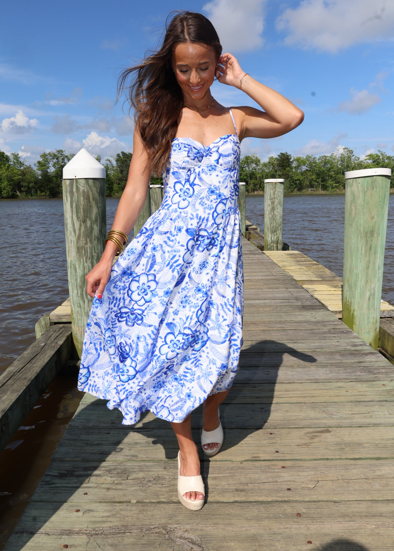 Woman in a blue floral dress standing on a wooden dock by a body of water.