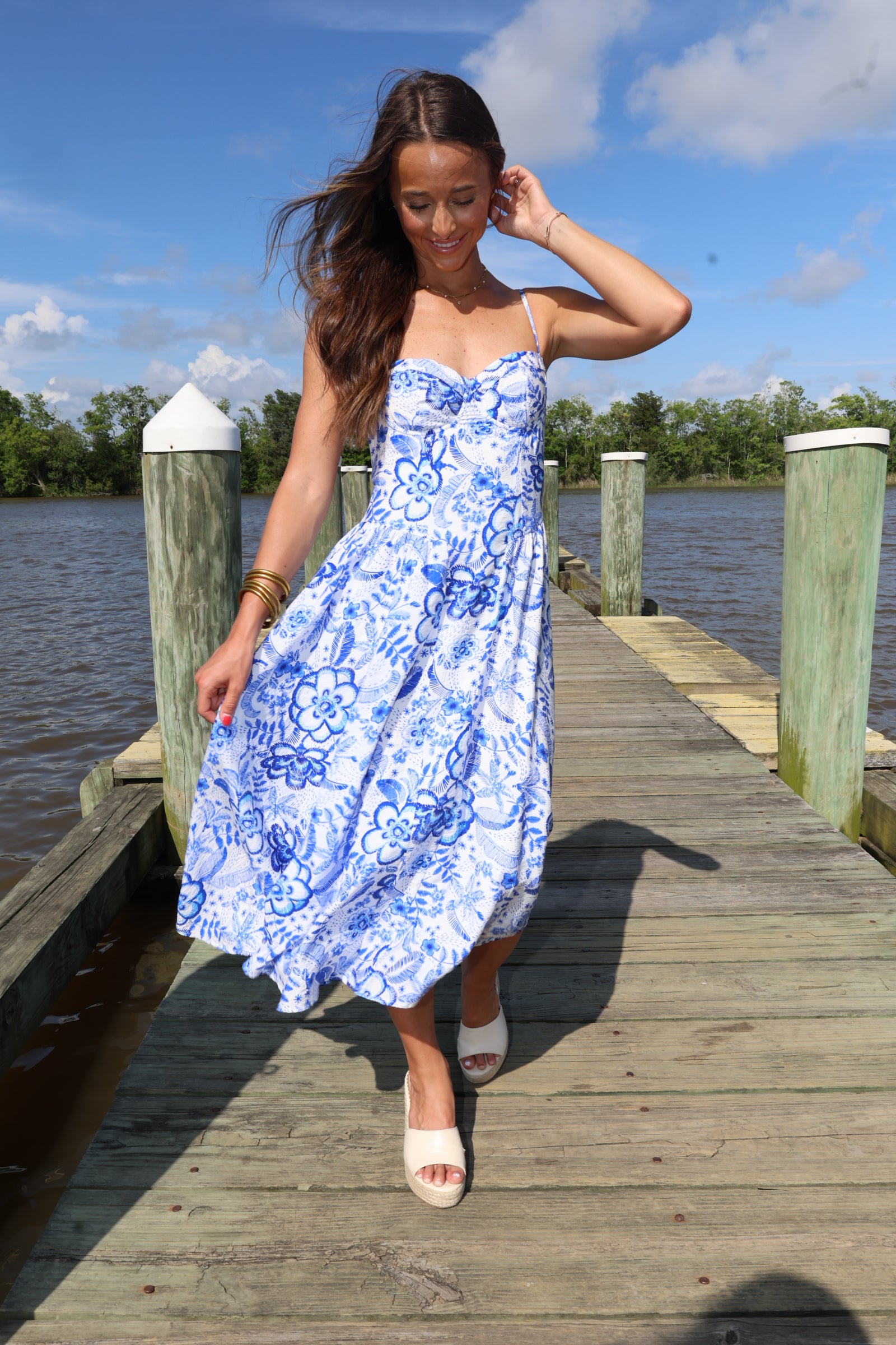 Woman in a blue floral dress standing on a wooden dock by a body of water.