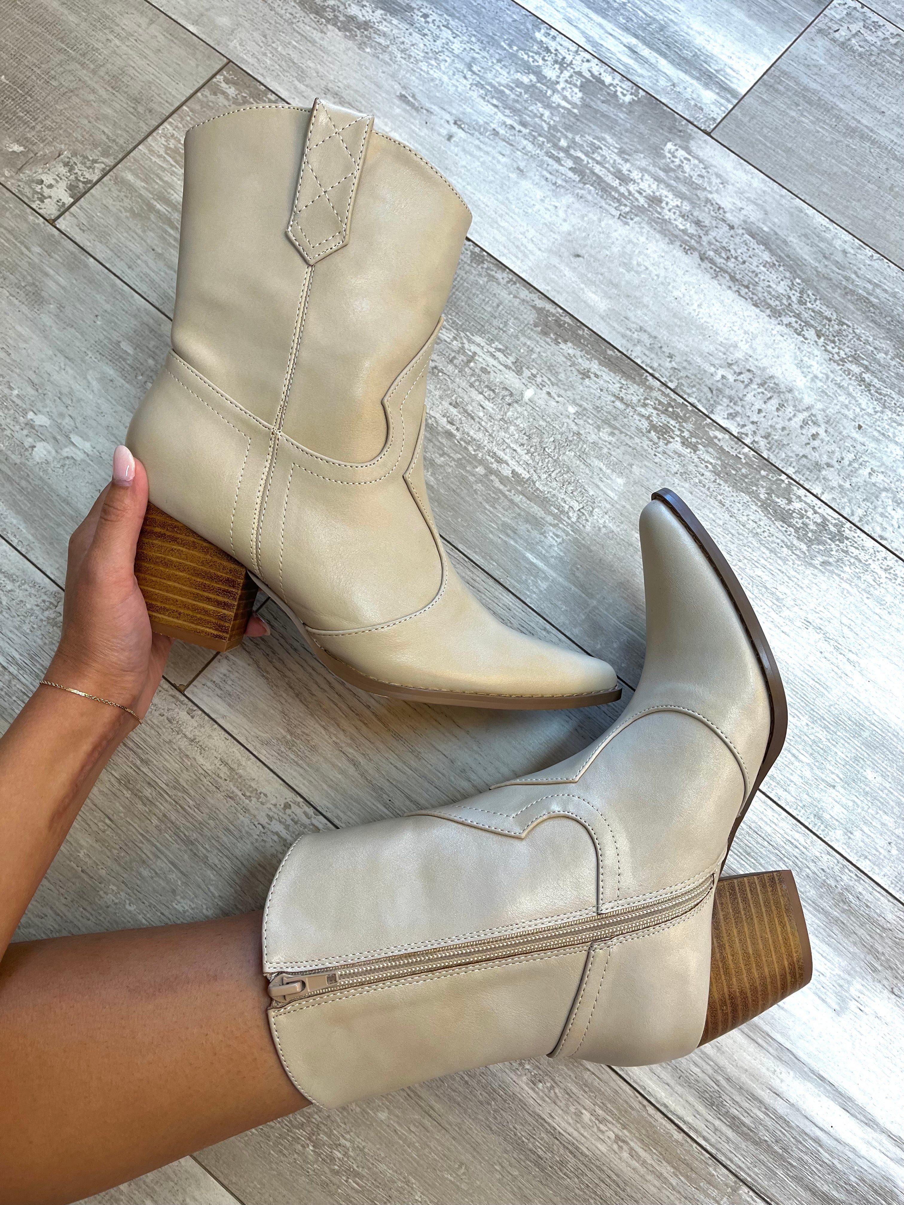 A pair of beige ankle boots with a block heel and side zipper, displayed on a person standing on a wooden floor.