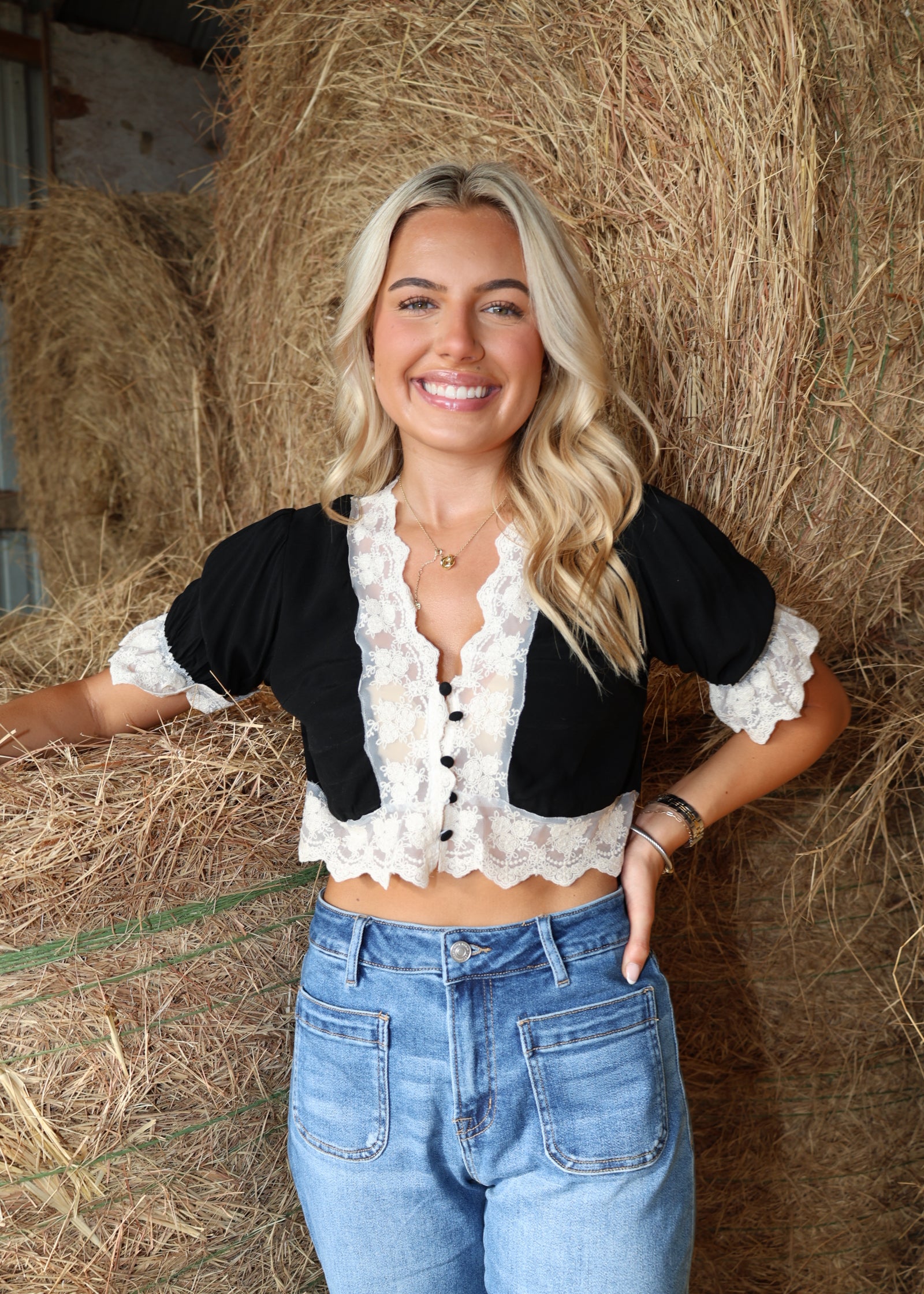 A smiling blonde woman stands in front of a large hay bale wearing a black cropped top with white lace trim and blue jeans.