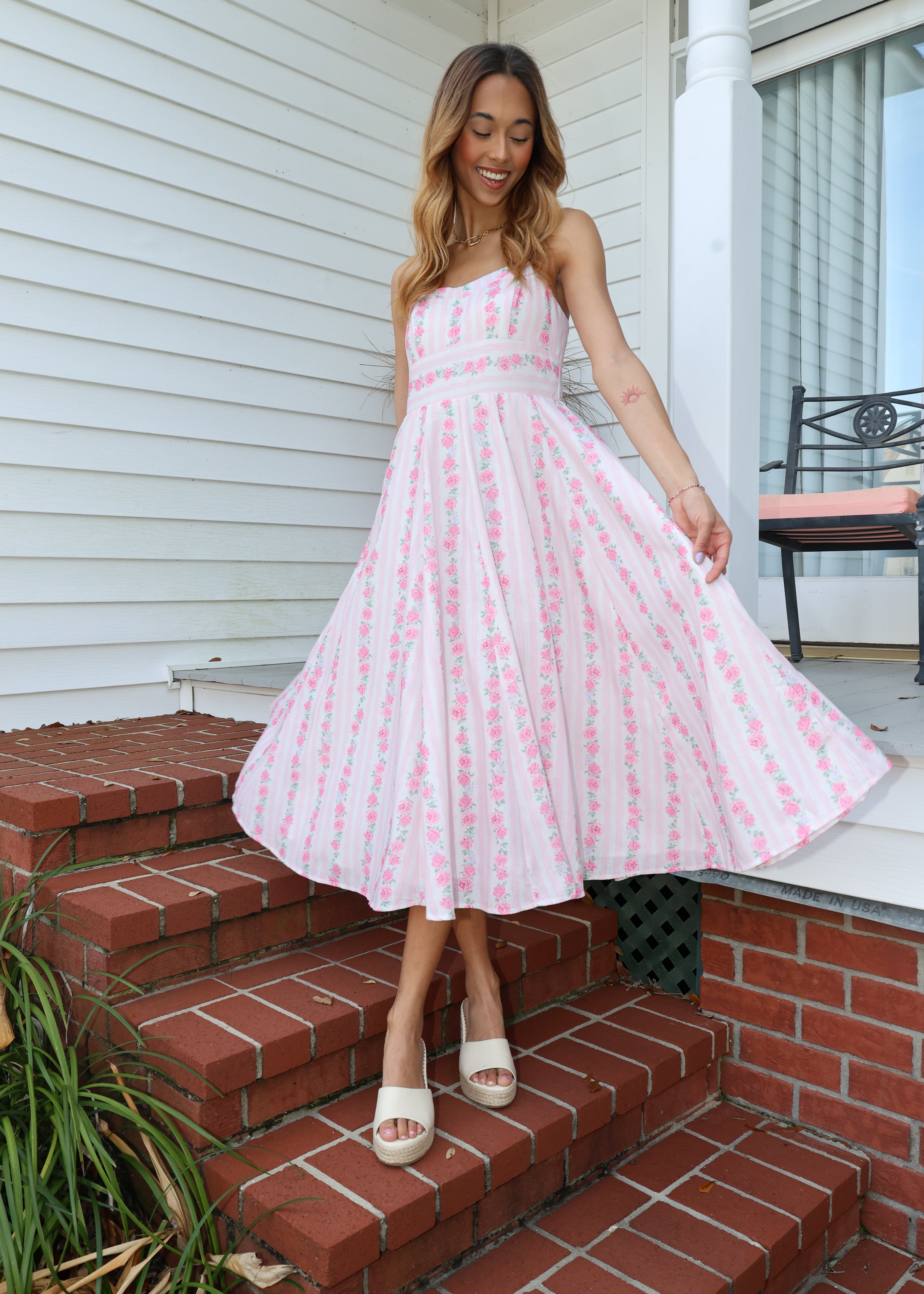 Woman in a pink dress standing on a porch with a brick wall and white door in the background.