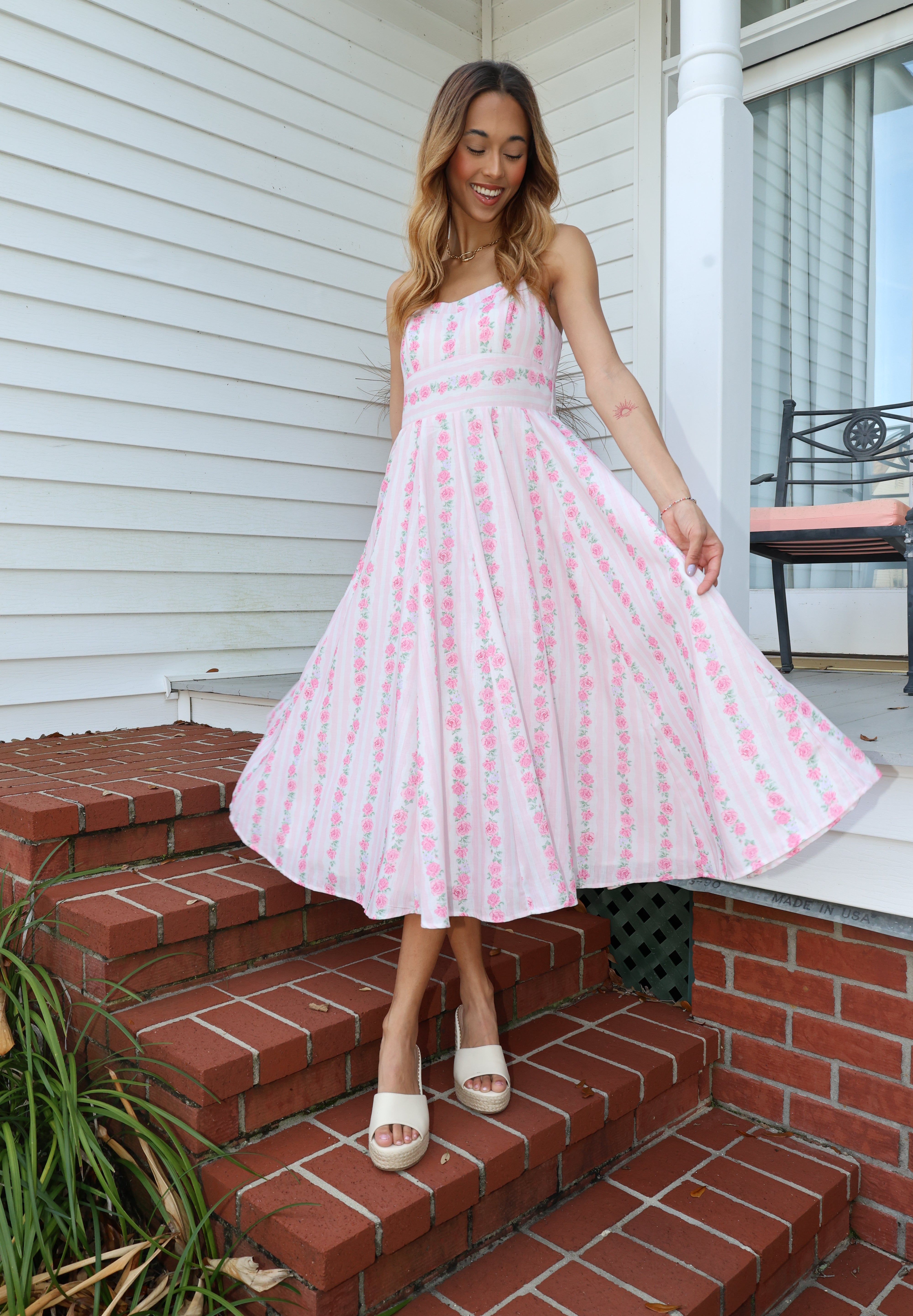 Woman in a pink dress standing on a porch with a brick wall and white door in the background.